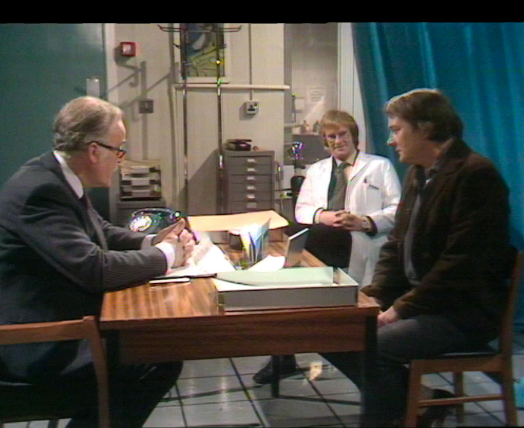 Film still from 'Follow the Yellow Brick Road'. Three men are in a clinical-looking office. One man in a suit sits at a desk with papers, facing a casually dressed man seated opposite him. A third man in a white lab coat sits nearby, observing. The room has medical equipment and storage cupboards.