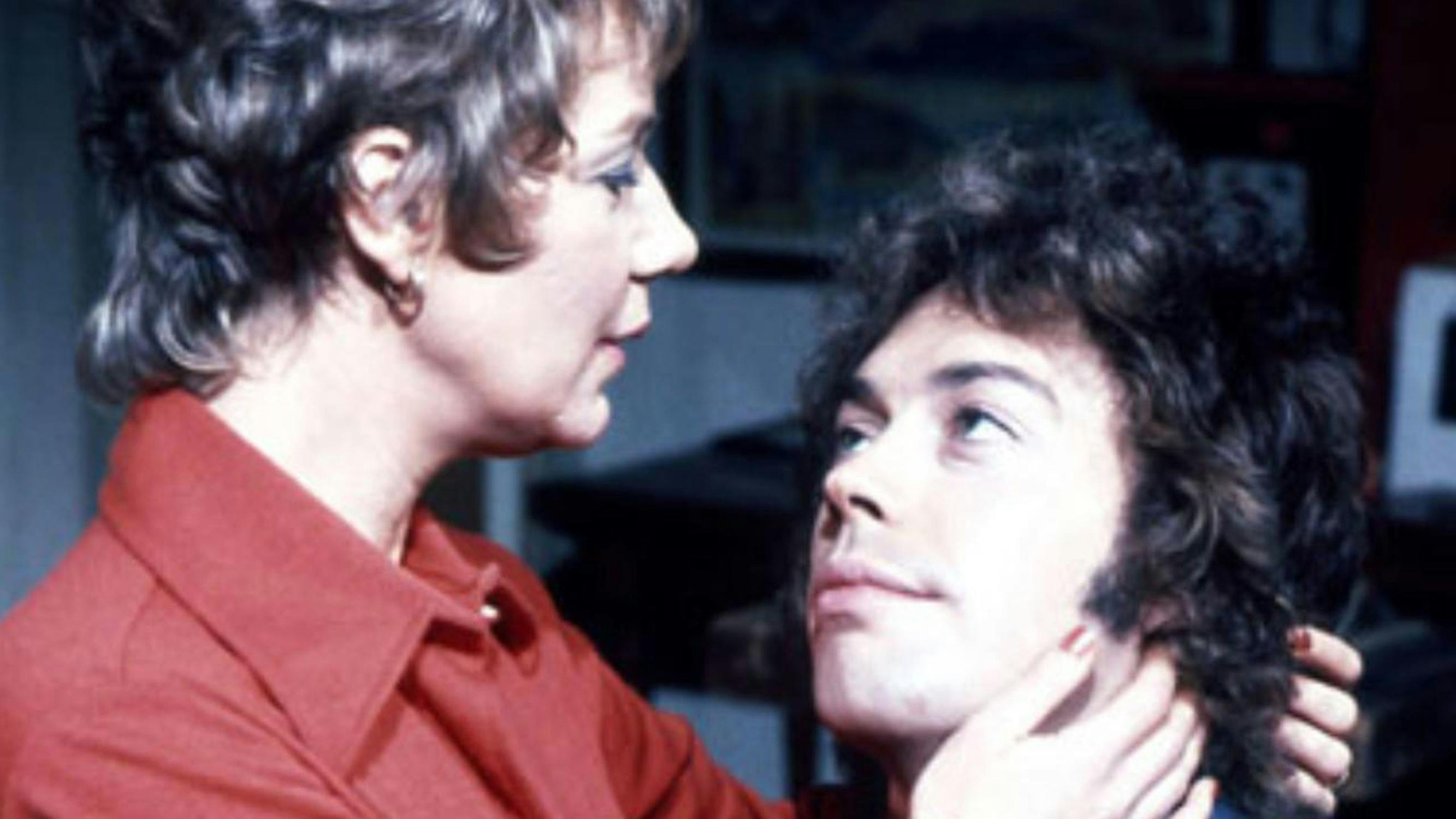 Film still from 'Schmoedipus'. A woman with short, light brown hair and a red collared shirt gently holds a younger man’s face, looking at him with concern. The man, with curly dark hair, looks up at her. The background is blurred, suggesting an indoor setting.