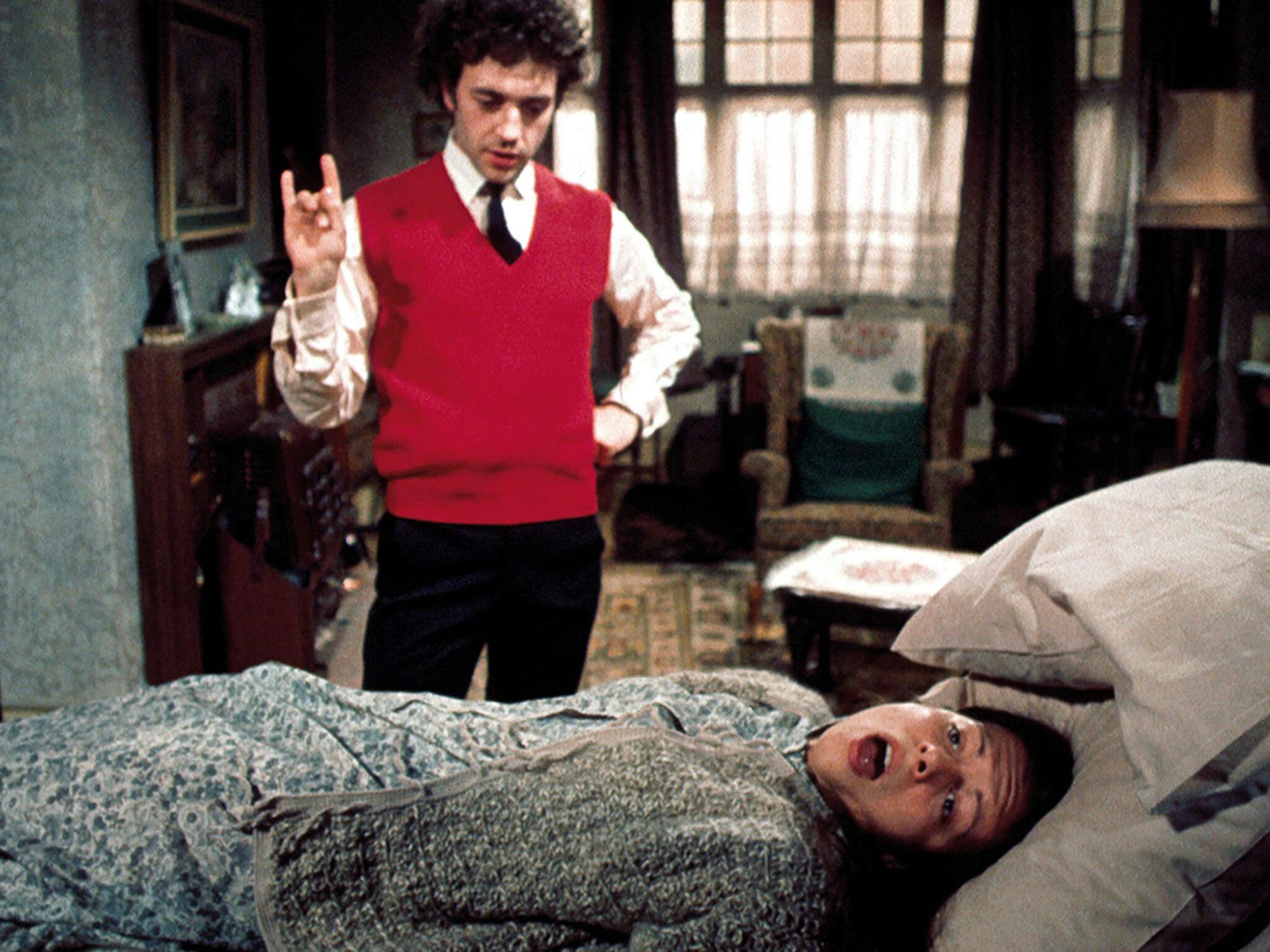 Film still from Brimstone and Treacle. A man in a red waistcoat stands with his hand raised, making a gesture, beside a woman lying on a bed with her mouth open in shock or distress. The scene occurs in a dimly lit, cluttered room with vintage furniture and patterned fabrics.