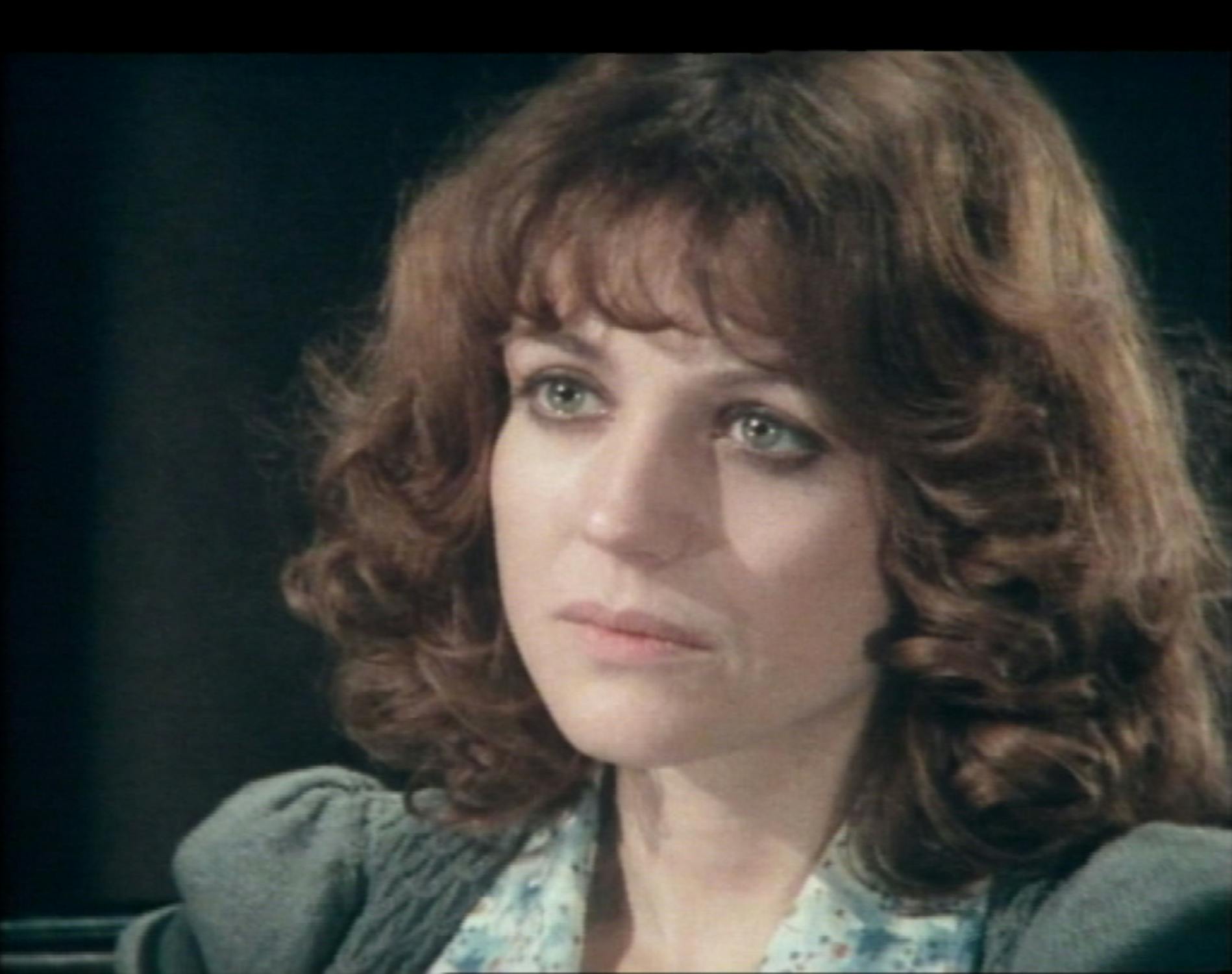 Film still from Double Dare. A woman with wavy, shoulder-length brown hair and green eyes looks thoughtfully ahead. She wears a patterned blouse under a grey jumper. Her expression is serious and contemplative, with soft lighting highlighting her face against a dark background.