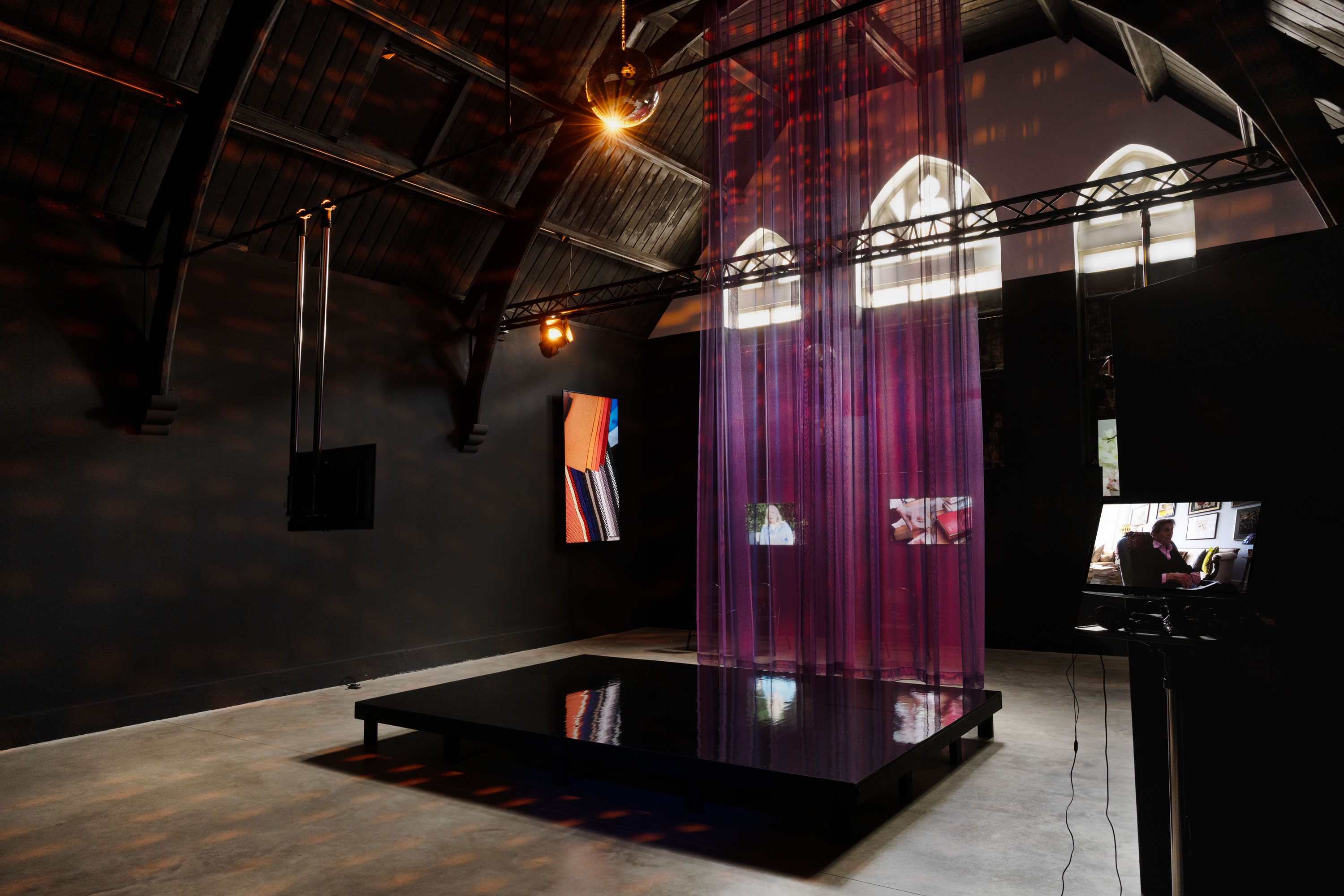 A dark art gallery with arched ceilings features a black reflective platform, vertical sheer purple curtains, spotlights, and video screens showing various abstract and figurative films. Three arched windows filter daylight into the space.