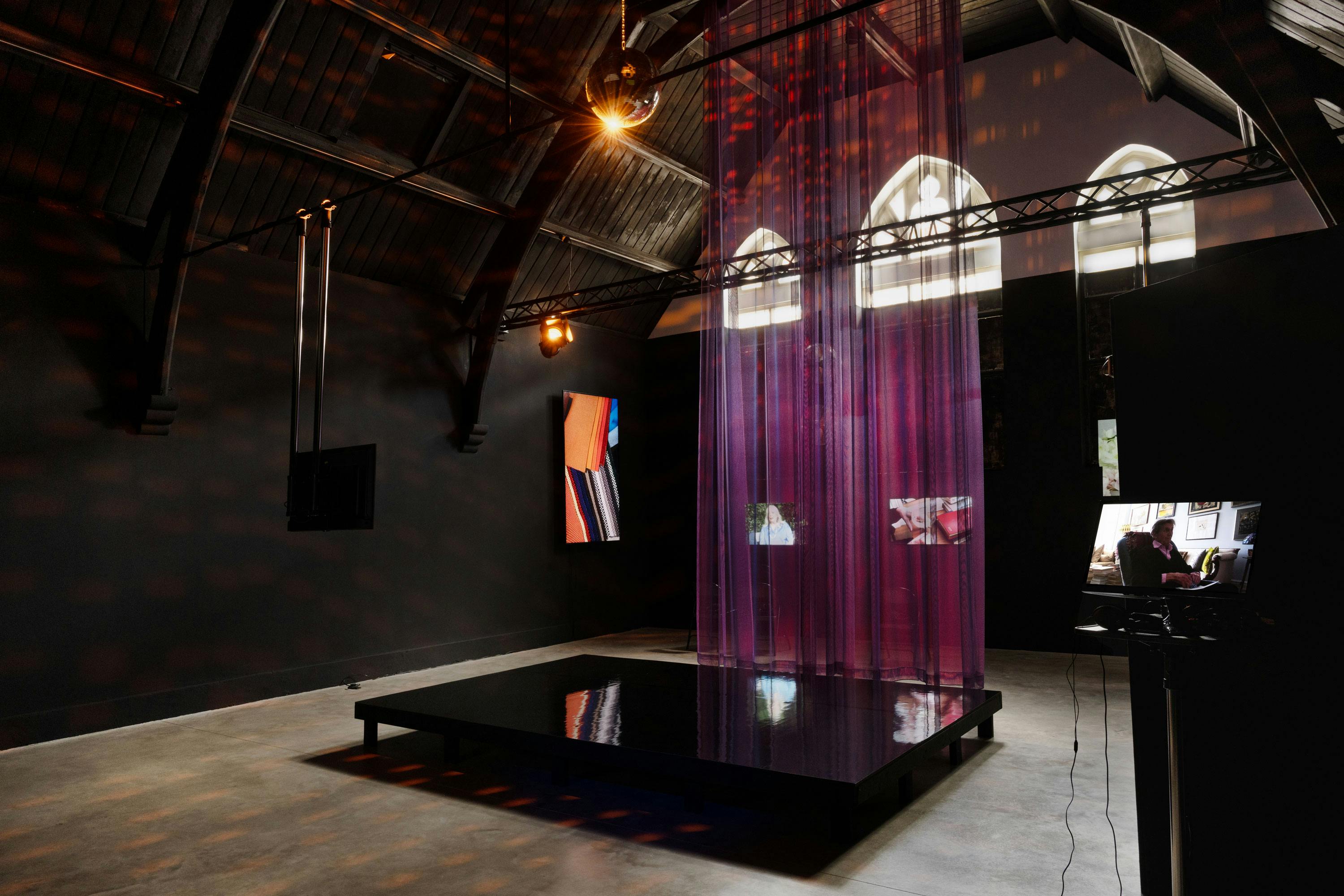 A dark art gallery with arched ceilings features a black reflective platform, vertical sheer purple curtains, spotlights, and video screens showing various abstract and figurative films. Three arched windows filter daylight into the space.