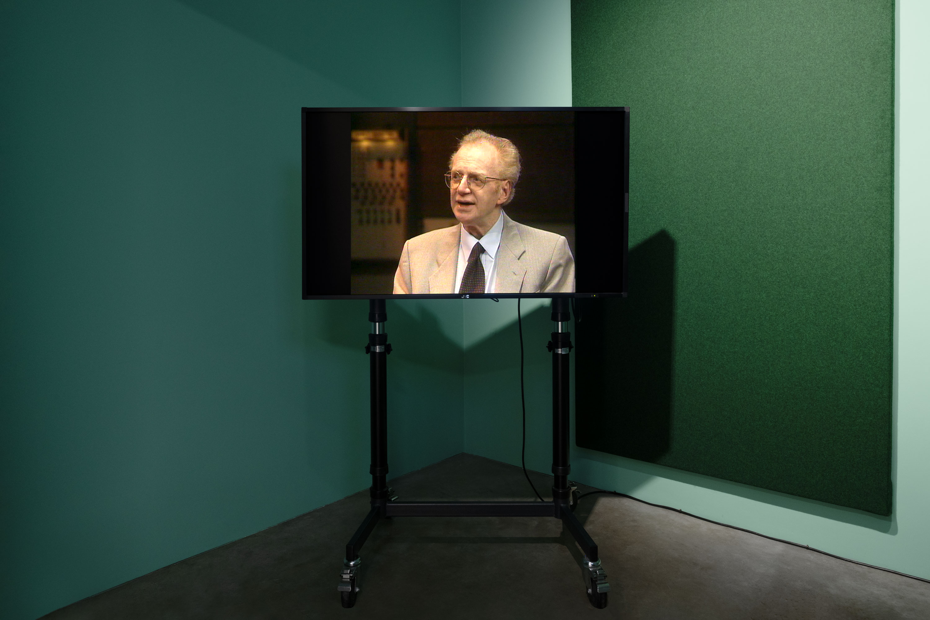 A television on a black wheeled stand displays an image of Dennis Potter wearing a beige suit, sitting and talking. The TV is in a corner of a green-walled room with dark flooring and an acoustic panel on one wall.