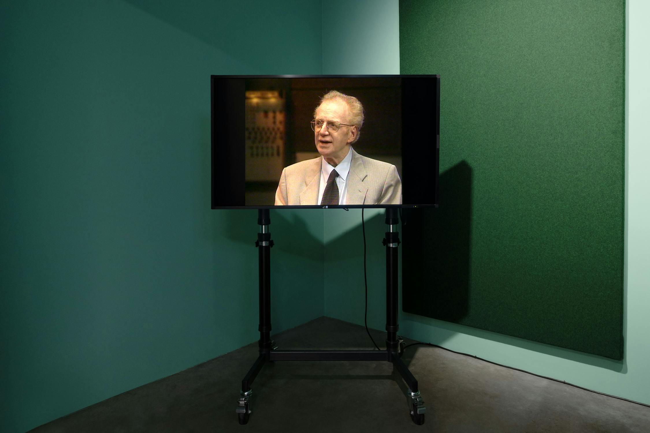 A television on a black wheeled stand displays an image of Dennis Potter wearing a beige suit, sitting and talking. The TV is in a corner of a green-walled room with dark flooring and an acoustic panel on one wall.