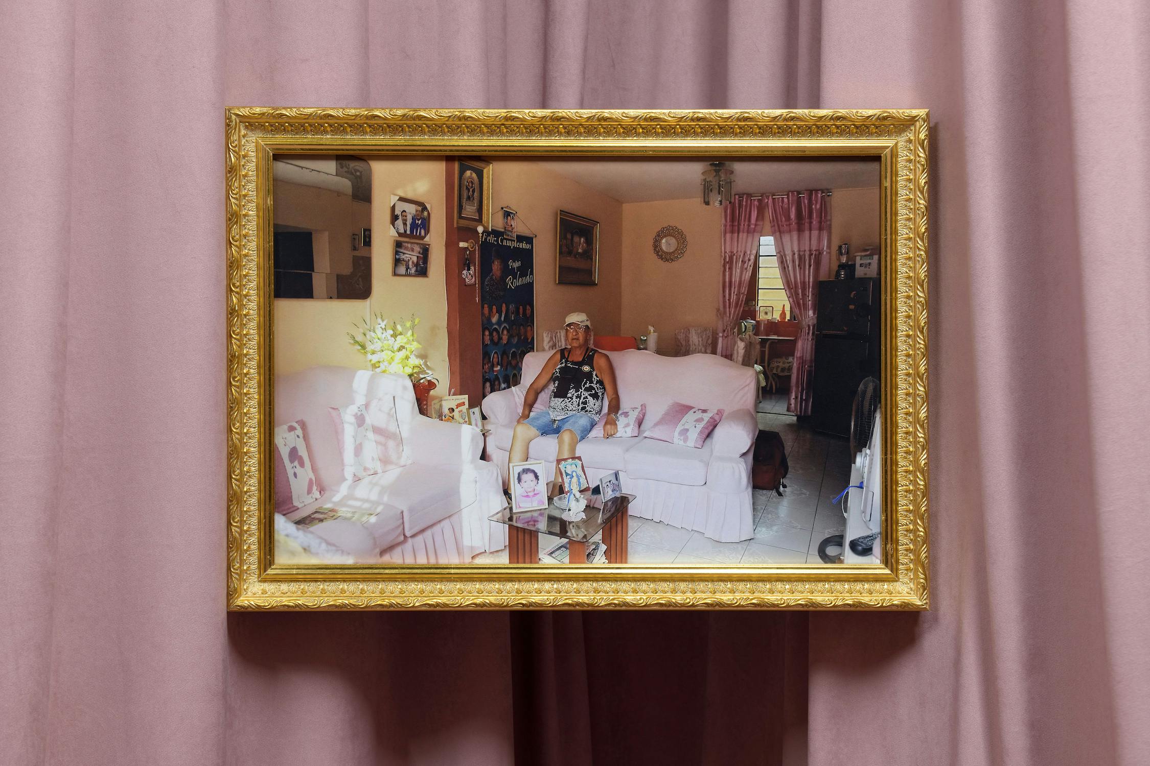 A framed photograph hangs on a mauve curtain. The photo shows an older woman with grey hair sitting on a white sofa in a cosy, pastel-coloured living room filled with family photos, decorations, and floral cushions. 