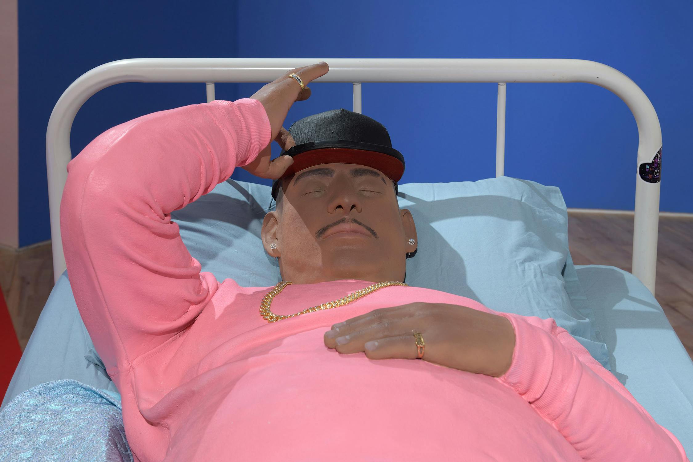 A sculpture of a man with a light brown skin tone lies on a bed in a pink sweatshirt, gold chain, and black cap, resting their left arm on their forehead. The background is blue, and the bed has light blue sheets and a simple white frame.