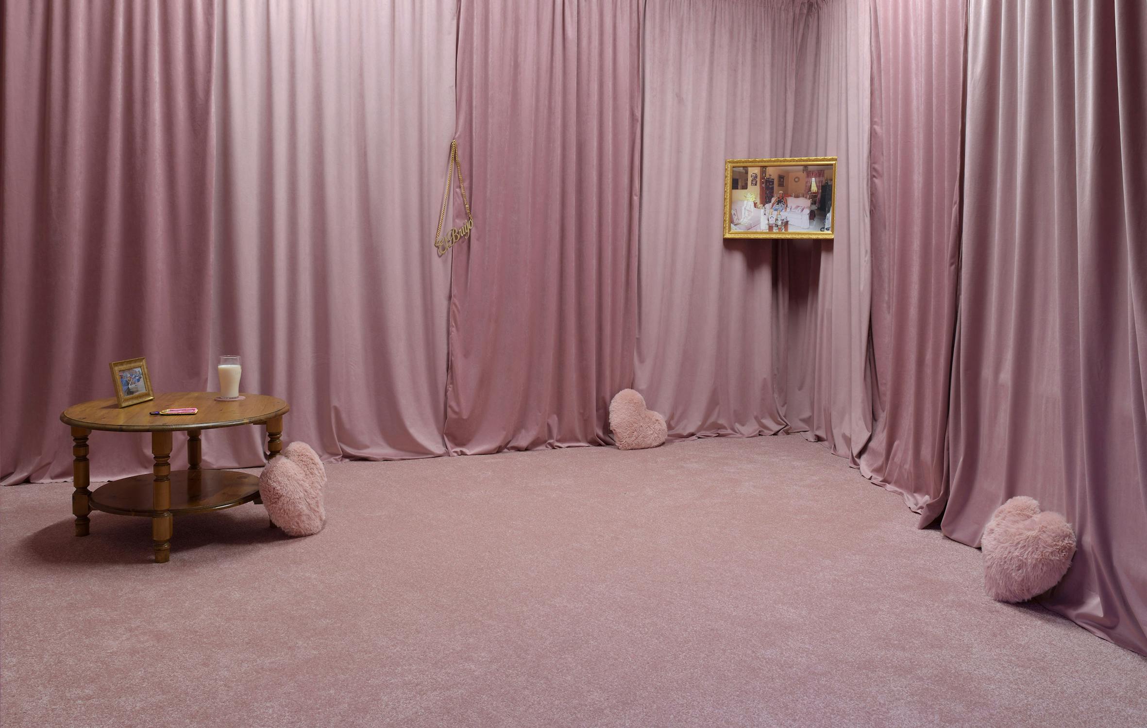 An art installation with floor-to-ceiling pink curtains and pink carpet, decorated with heart-shaped cushions. A round wooden table holds a framed photo, a candle, and a red notebook. A gold-framed mirror and picture are on the curtain-covered walls.