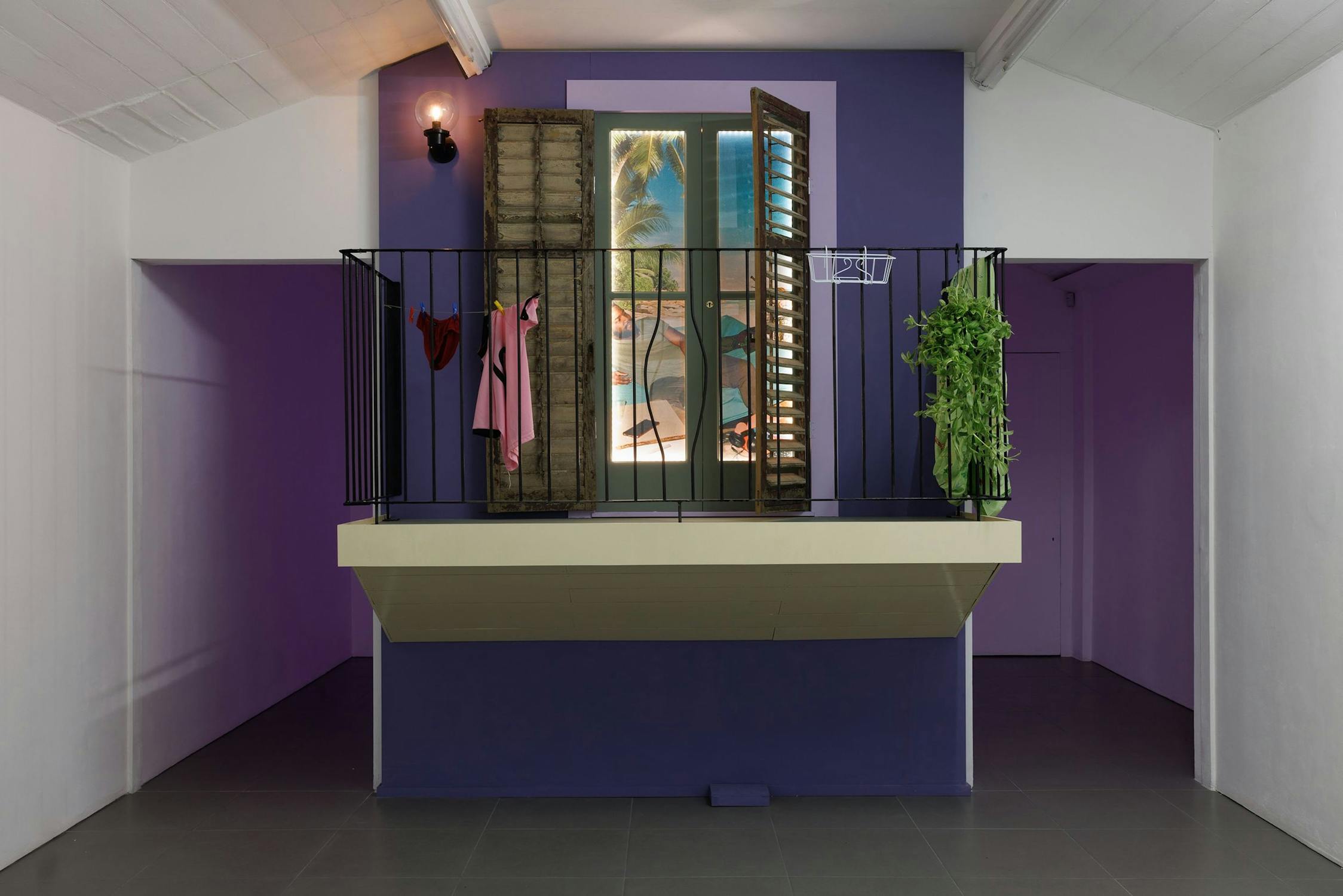 A small indoor balcony with black railings juts from a purple wall. Clothes are draped over the railing, a green hanging plant is on one side. Open wooden shutters reveal a window with a tropical beach scene outside. The ceiling and walls are white.
