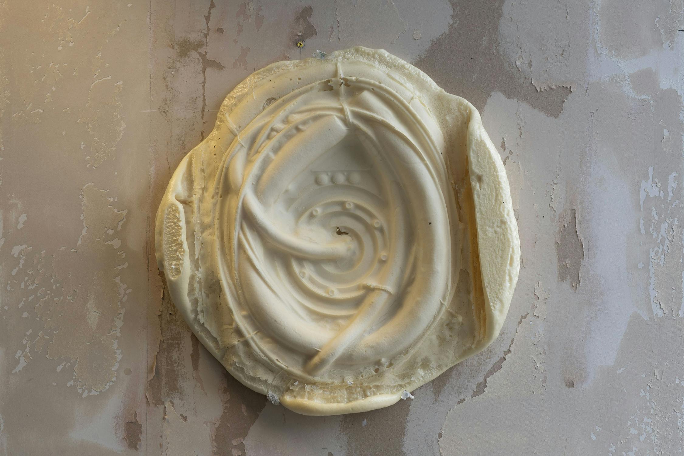 A round, beige plaster or clay relief by Chaney Diao with abstract curved and spiral patterns hangs on a textured, weathered wall with uneven paint and patches of grey and taupe. The relief has raised edges and intricate lines forming a central swirling design.