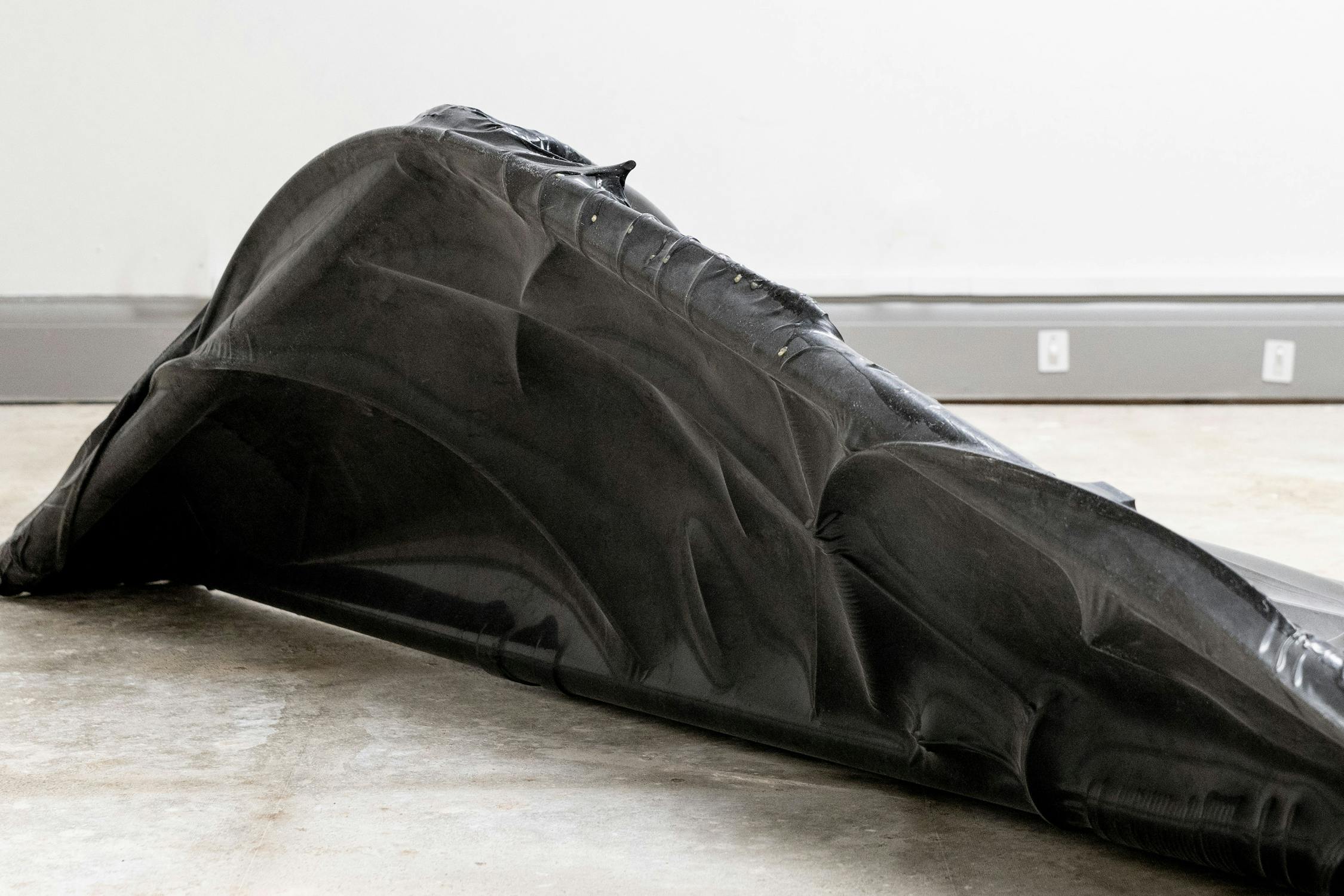 A large, black latex sculpture resembling stretched, crumpled fabric or rubber lies on a concrete floor against a plain white wall. The material forms angular and curved shapes, creating an abstract, organic appearance.