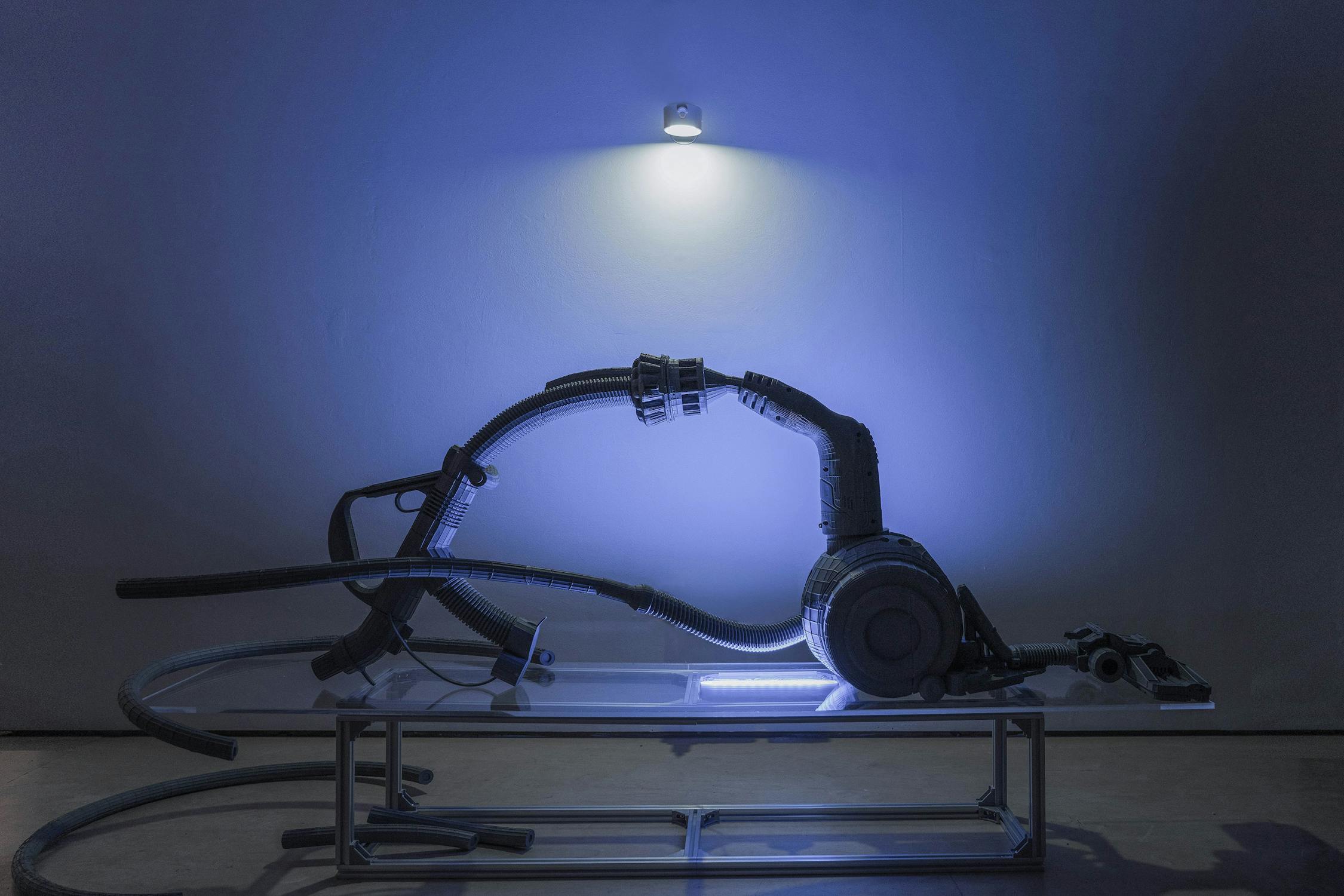 A metallic, futuristic sculpture made from the distorted and warped parts of a vacuum cleaner sits on a glass-topped table, illuminated by blue light from above, casting shadows on the wall and floor in a minimalist, modern setting.
