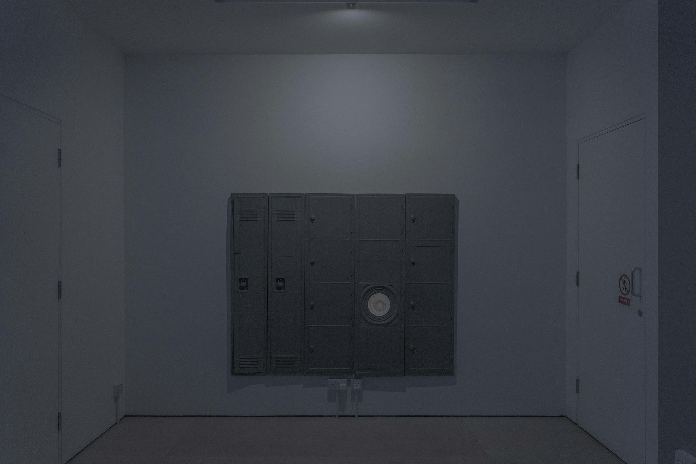 A dimly lit corridor with pale walls features two closed white doors—one on each side—with an art work resembling dark lockers mounted to the back wall. A round speaker or vent is fitted in the centre locker. A “No Smoking” sign is visible on the right door.