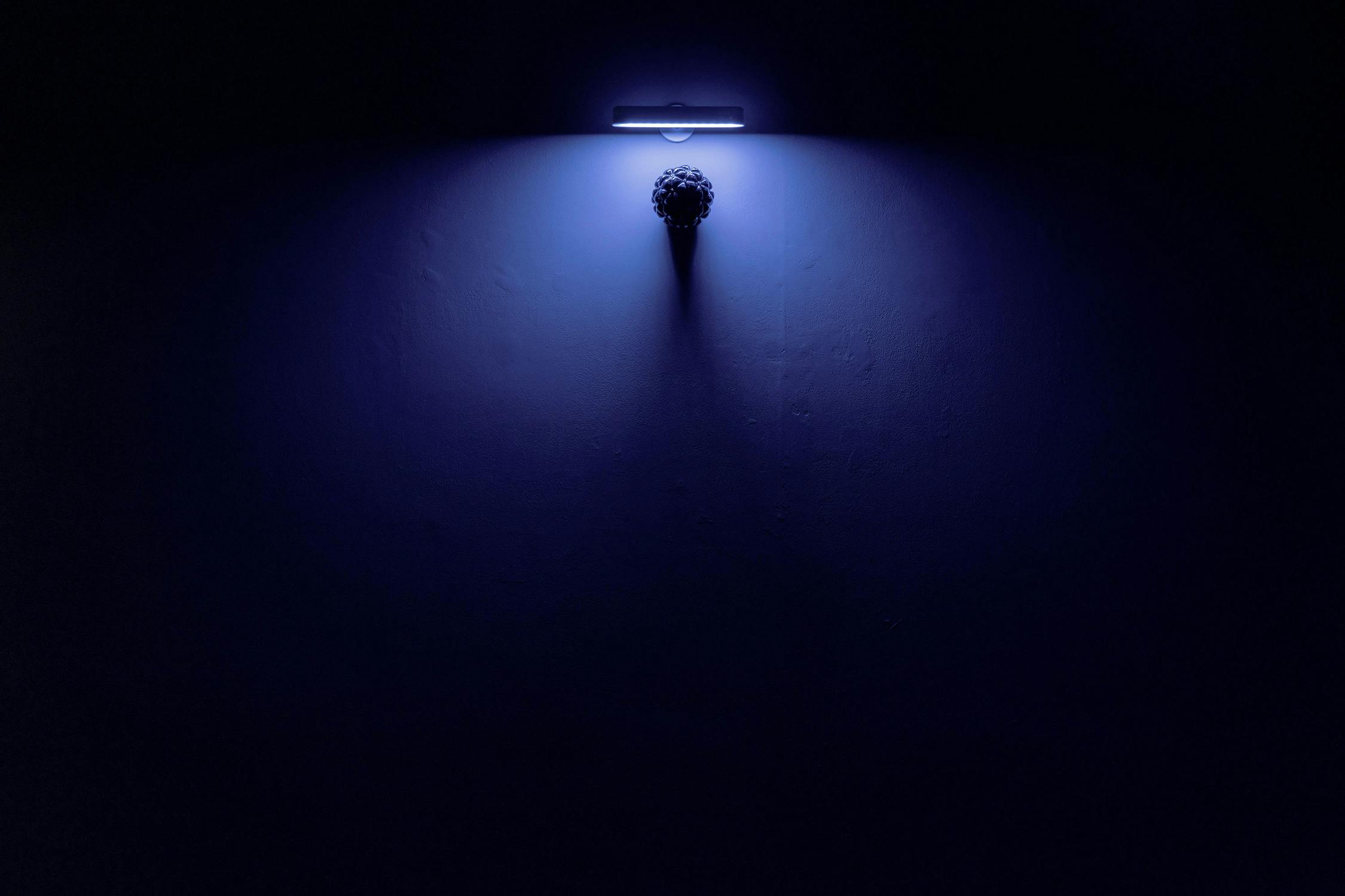 A single overhead light fitting casts a dim, bluish glow on a dark, empty wall. The rough texture of the wall is faintly visible in the illuminated area, which is otherwise surrounded by deep shadows and darkness.