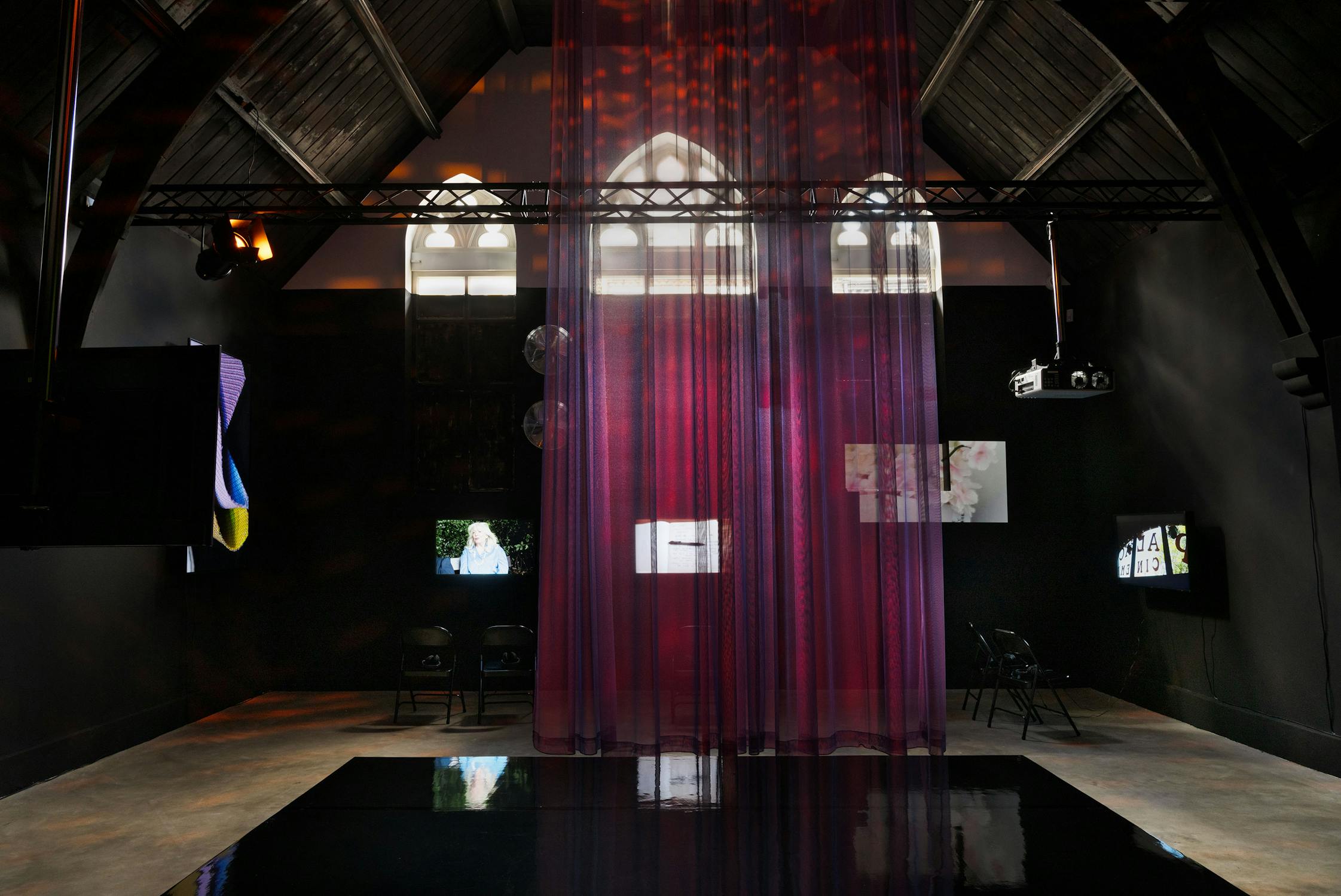 A dimly lit room with high vaulted ceilings, stained glass windows, and translucent purple curtains hanging from above. Black chairs sit near the walls, and several screens display images on the dark walls. The floor is reflective, adding to the moody ambience.