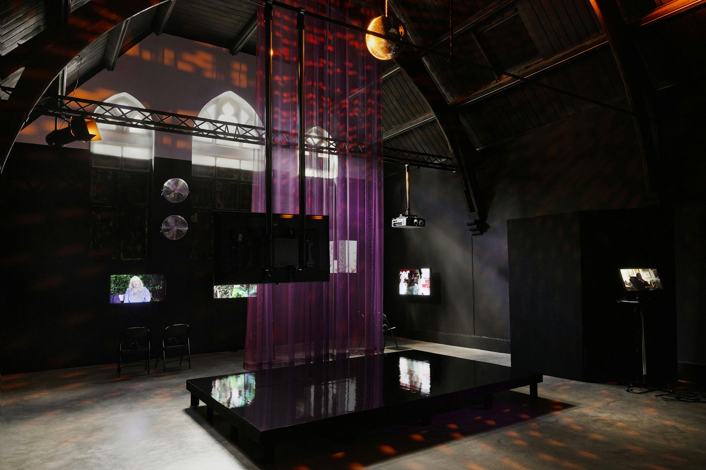 A dimly lit room with arched ceilings features a shiny black platform, sheer purple curtains hanging from above, wall-mounted screens showing images, industrial lights, and stained-glass-style windows casting coloured patterns onto the walls and floor.