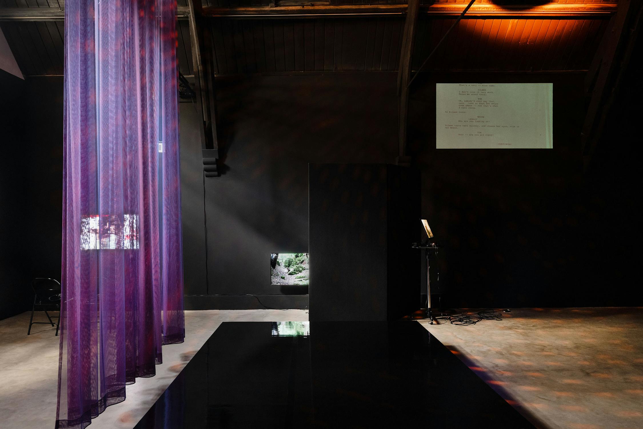A modern, dimly lit gallery features sheer purple curtains, a reflective black platform, and textured concrete floors. A small window in the black wall reveals greenery. A projected white screen with text appears near the ceiling above a small desk and equipment stand.