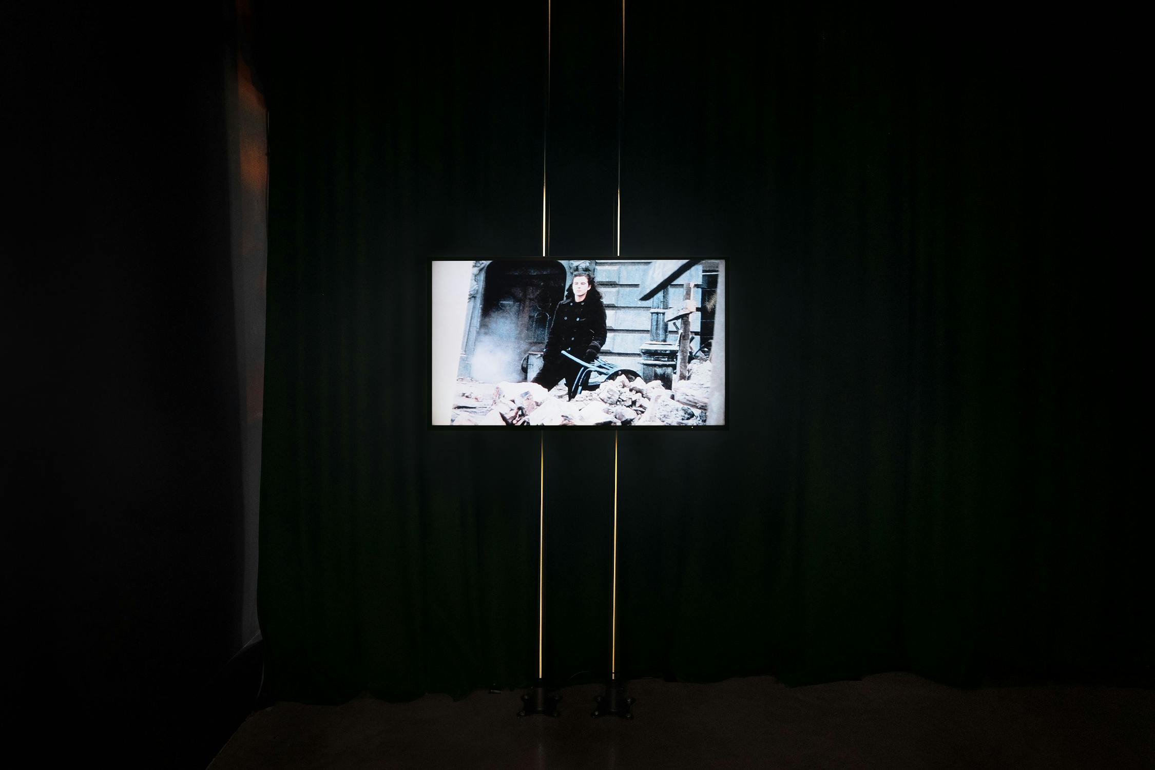 A television screen in a dark room displays a woman sitting among rubble. The screen is mounted against dark green curtains, with two vertical yellow light strips behind it. The rest of the room is dimly lit, emphasising the screen's content.