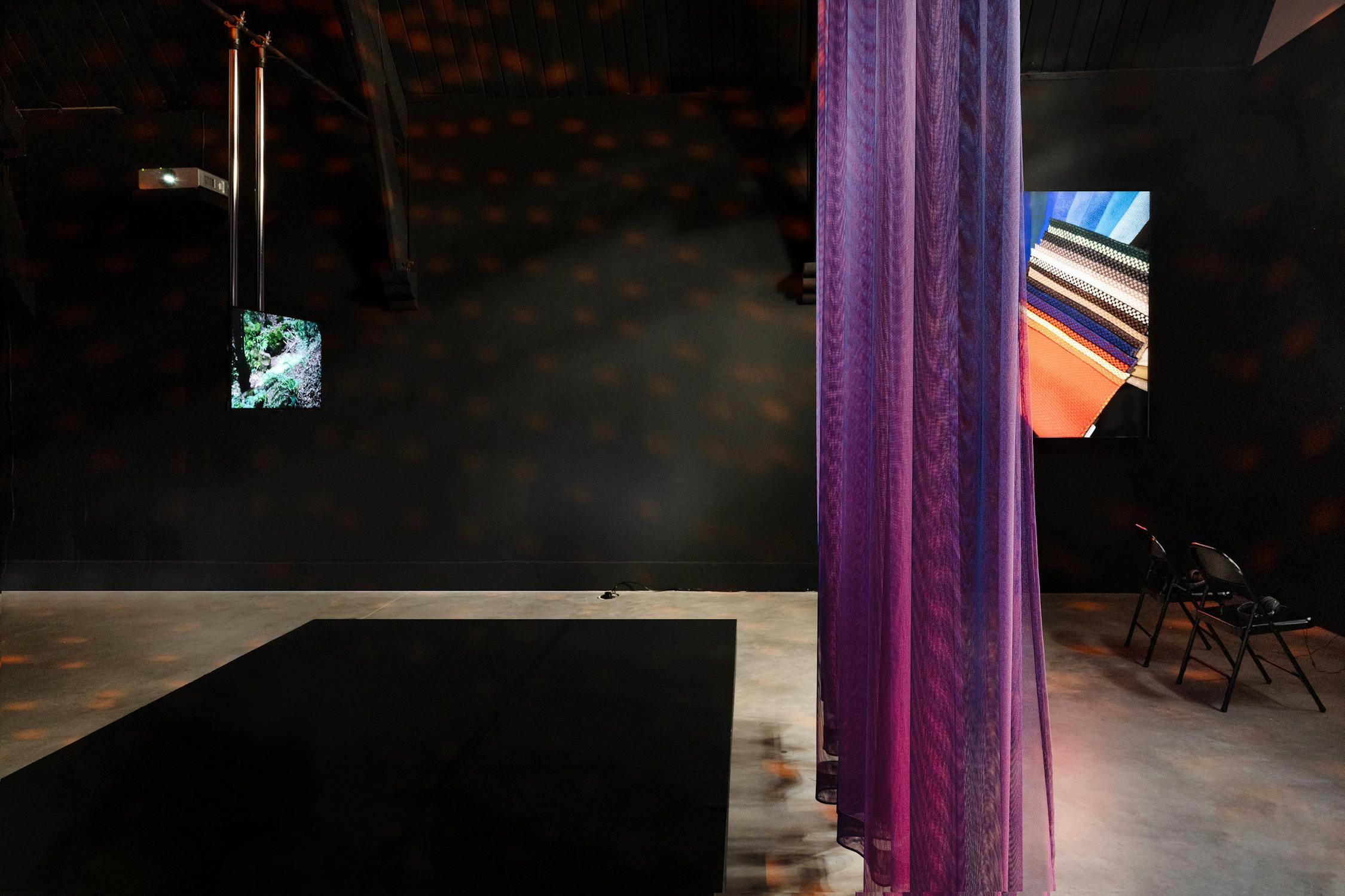 A dimly lit room features purple sheer curtains hanging in the foreground, two digital screens on the dark back wall—one showing a landscape, another abstract colours—and two black chairs arranged near the screens. Light spots dot the black walls and ceiling.