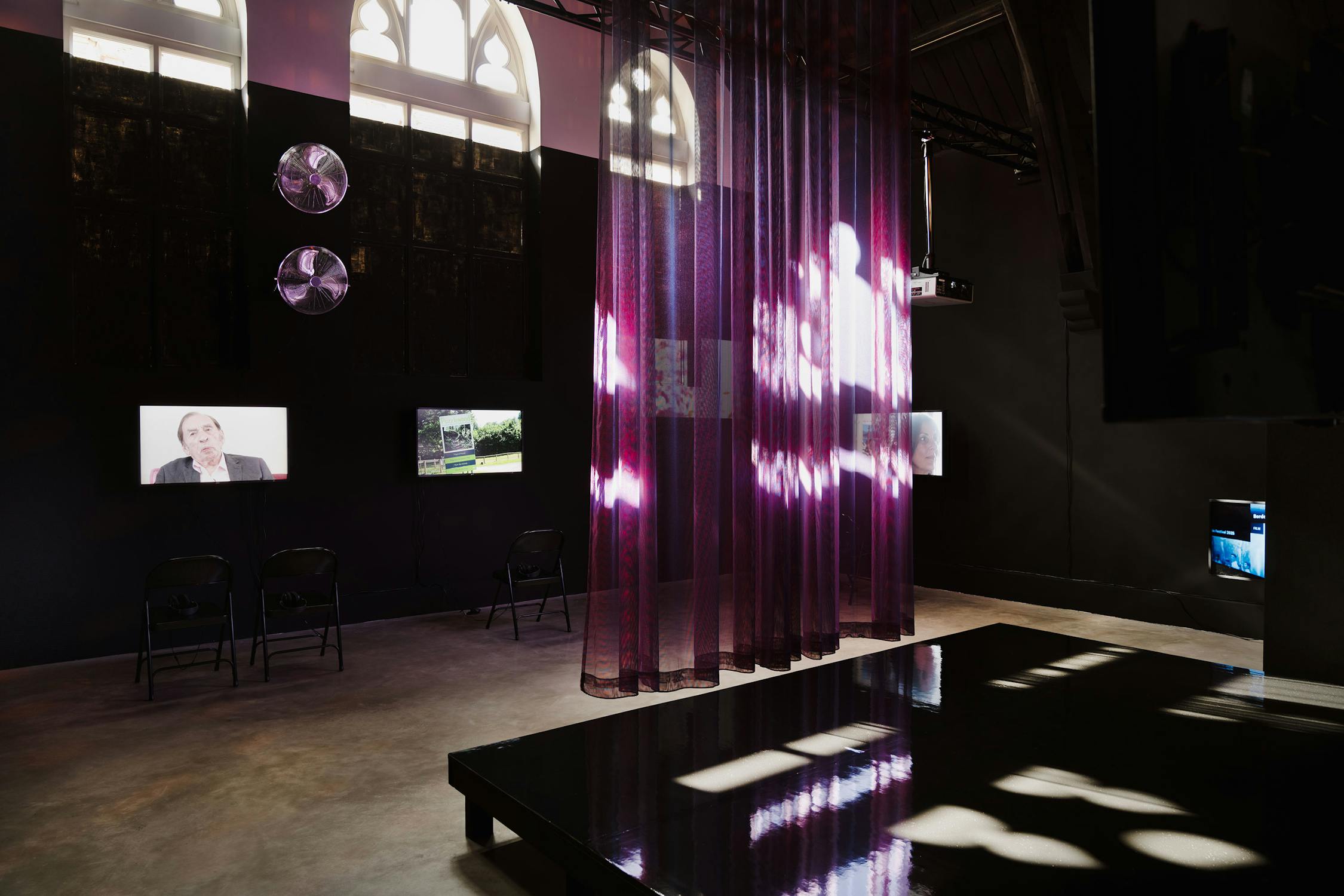 A dimly lit art installation features translucent purple curtains with light patterns, several video screens on black walls, two black folding chairs, reflective surfaces, and arched windows casting natural light into the space.