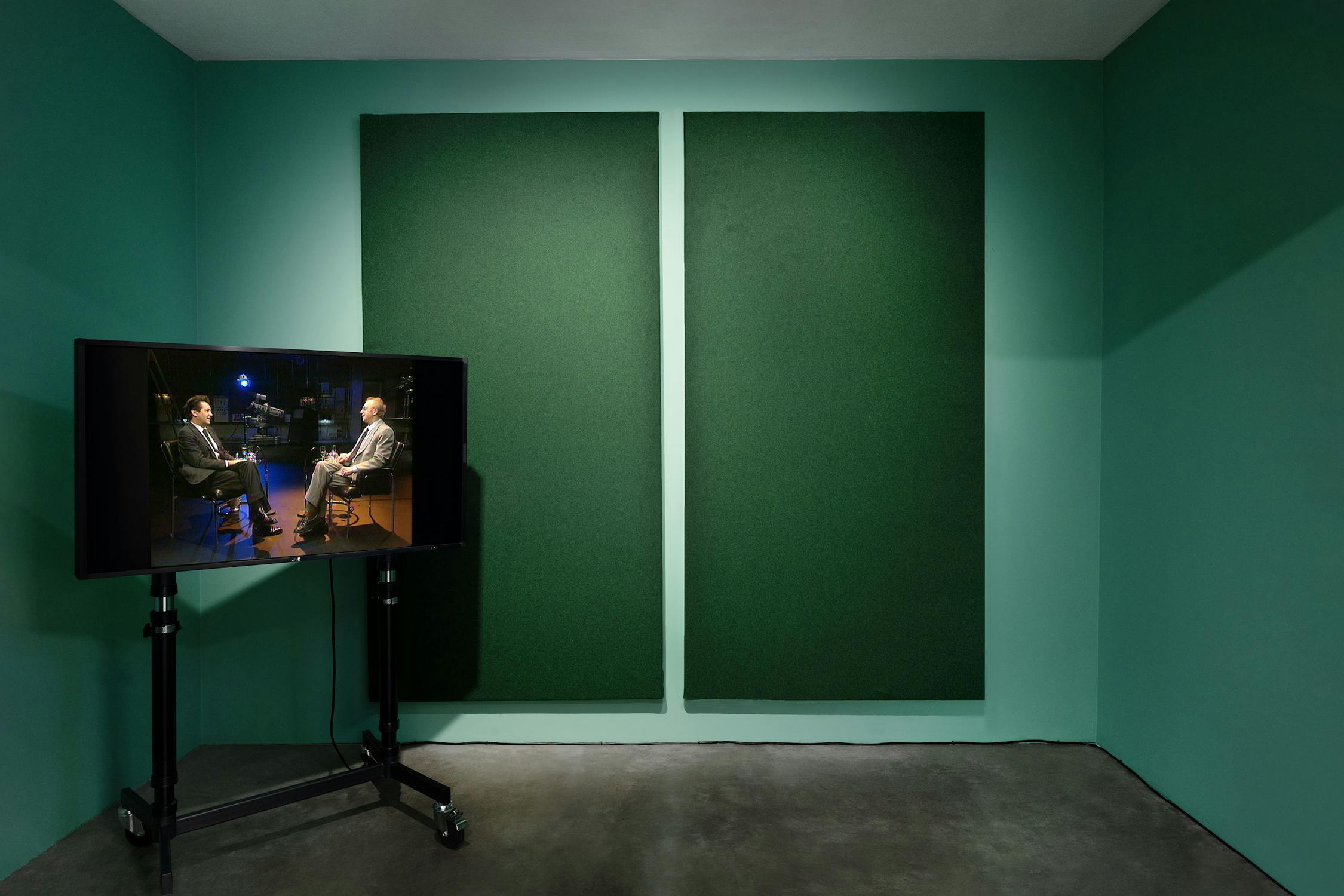 A small green-walled room contains a TV on a stand, displaying Dennis Potter and Melvyn Bragg sitting and talking. The TV faces the room’s empty space. Two large dark green sound panels are mounted on the far wall above a concrete floor.