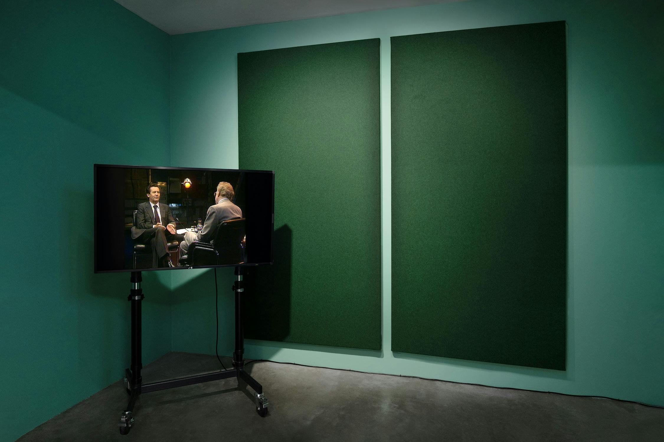 A television on a wheeled stand displays two people in suits having a conversation. The TV is in a minimalist room with teal walls, grey floor, and two large dark green panels mounted on the wall behind the screen.