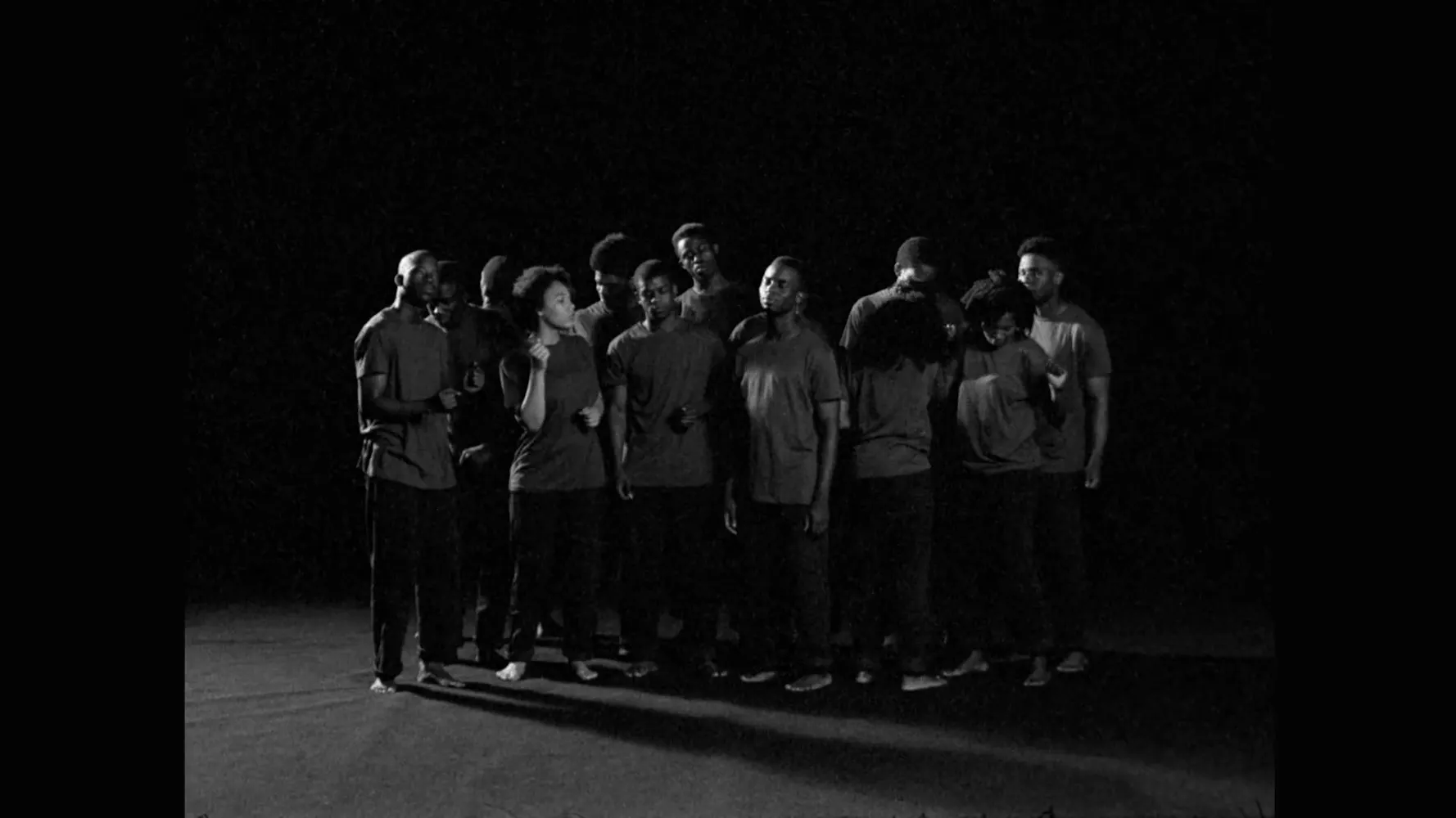 A group of about fifteen people stands closely together in a dimly lit, black-and-white setting. They wear casual, dark clothing and are clustered in the centre of the frame, surrounded by darkness, creating a dramatic and focused visual effect.