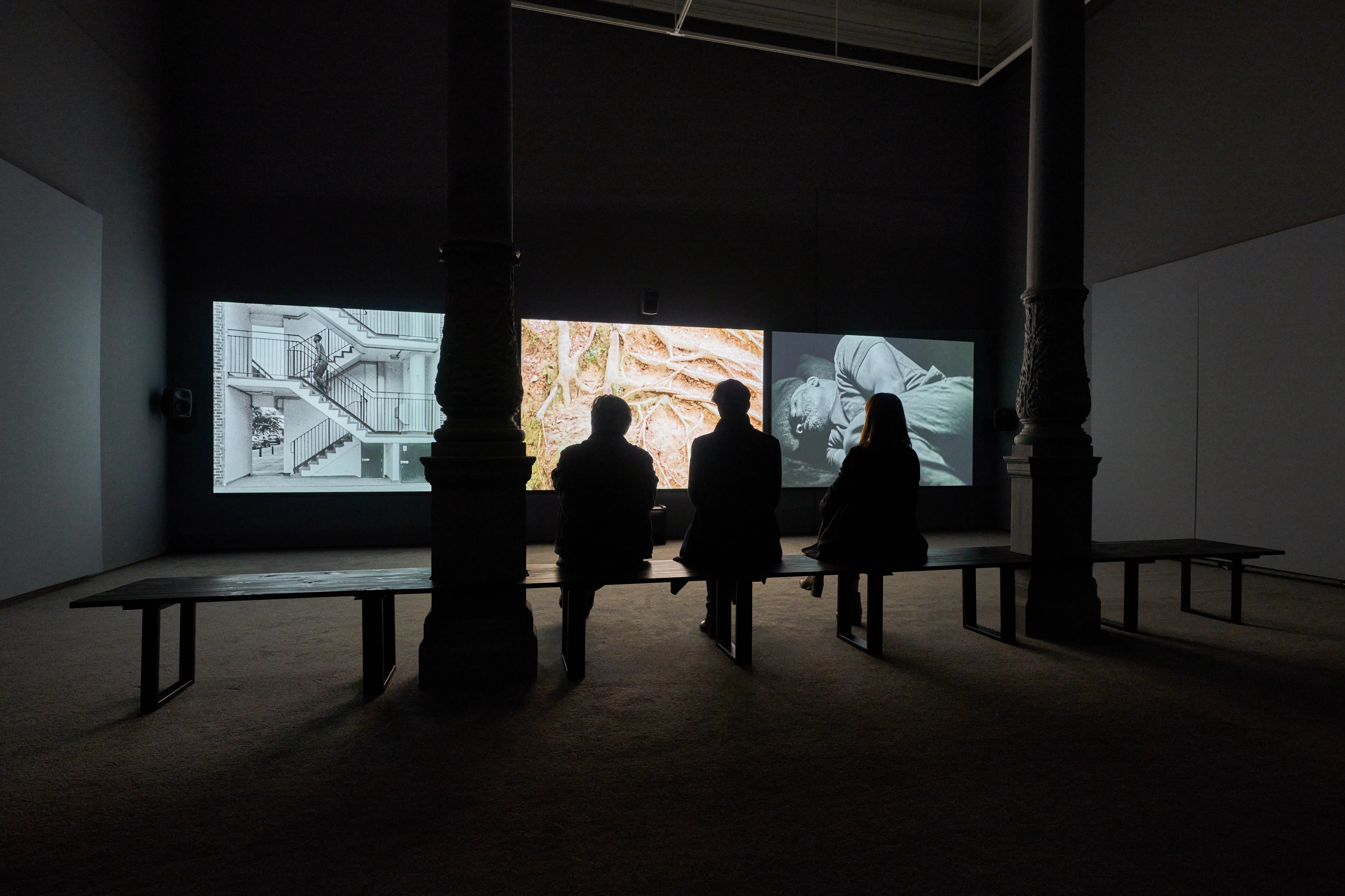 Three people sit on a bench in a dark gallery, facing three large screens showing black-and-white and colour images, including architectural details, tree roots, and a person lying down. Two columns frame the screens, creating a dramatic atmosphere.