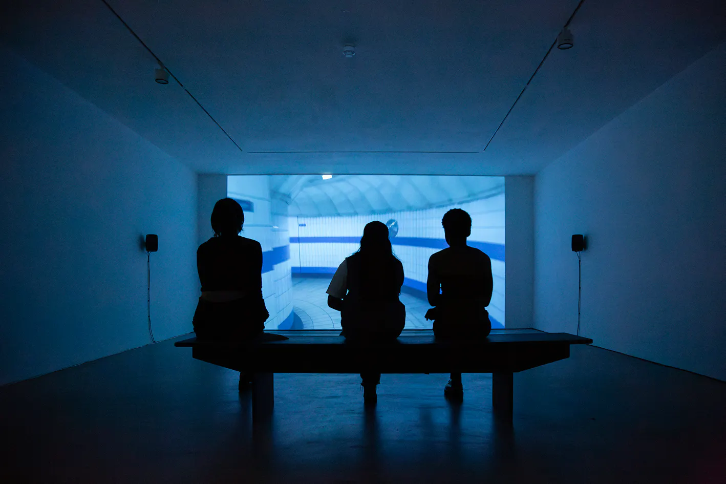 Three people sit on a bench in a dark room, silhouetted against a large, brightly lit screen showing a blue and white abstract or architectural scene. The minimalist room has white walls, ceiling lights, and small speakers mounted on either side of the screen.