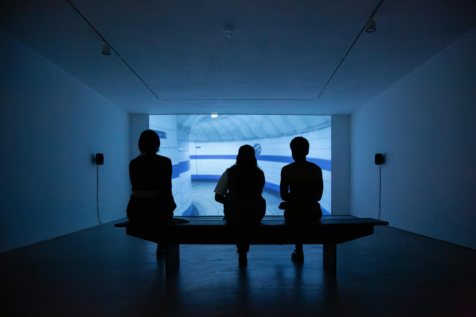 Three people sit on a bench in a dark room, silhouetted against a large, brightly lit screen showing a blue and white abstract or architectural scene. The minimalist room has white walls, ceiling lights, and small speakers mounted on either side of the screen.