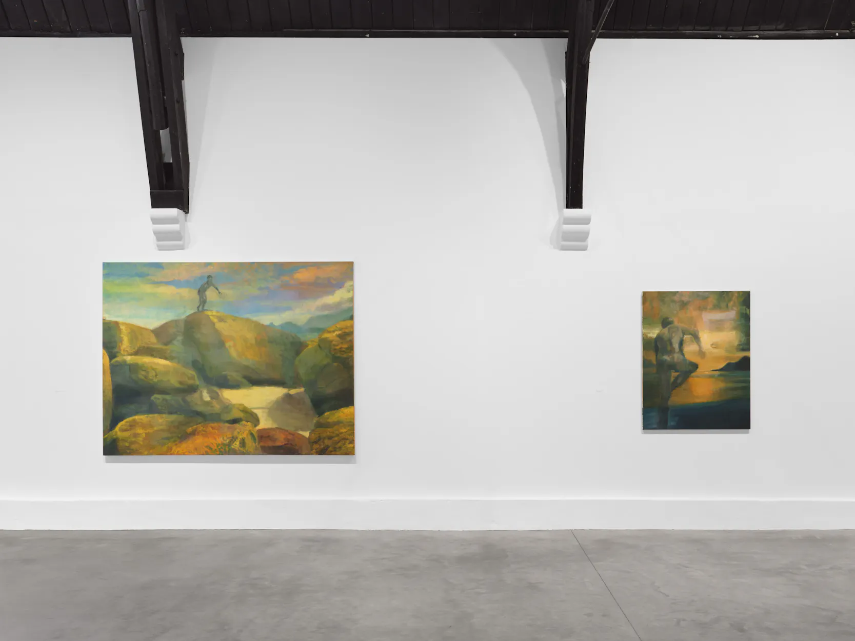 An art gallery wall displays two landscape paintings: one large painting on the left shows a figure atop colourful rocky terrain under a vivid sky, while the smaller painting on the right depicts a lone person near water with soft, warm lighting.