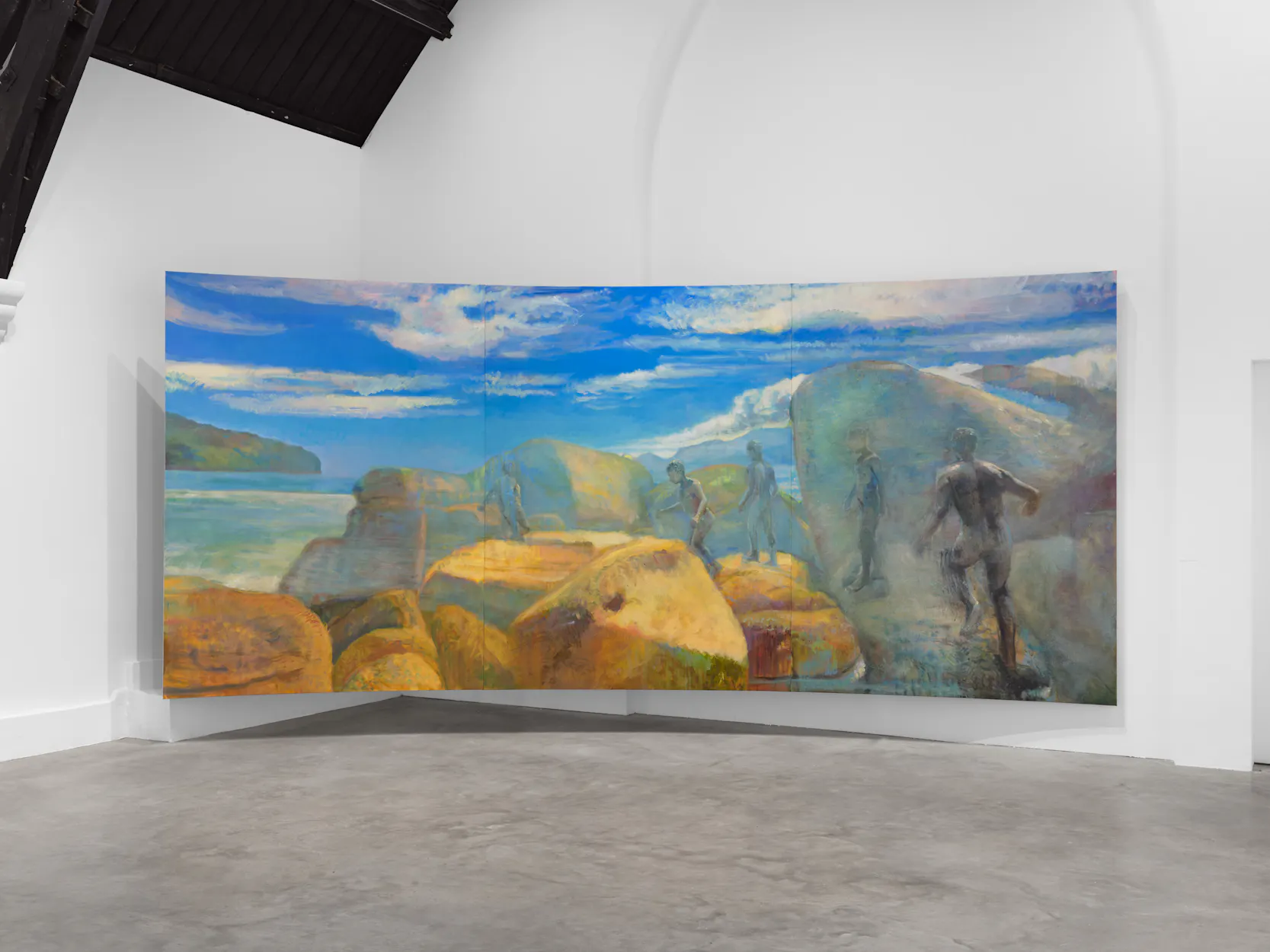 A large curved painting in a white gallery shows rocky terrain by the sea, with cloudy blue skies above. Faint, ghost-like human figures appear to walk among the yellow-orange rocks, blending partially into the landscape.