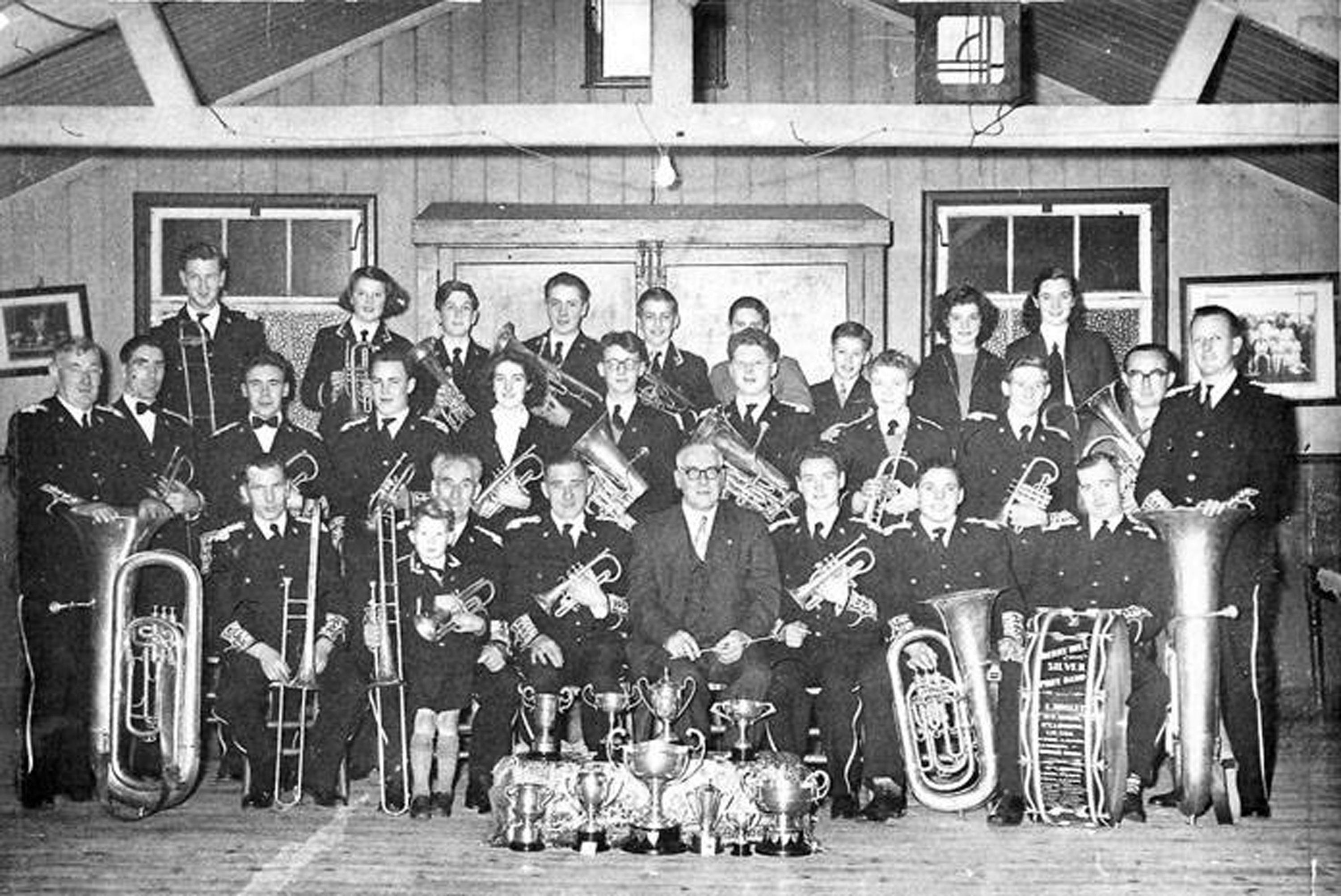 A black-and-white photo shows a large brass band of men, women, and children in uniforms posing indoors. They hold various brass instruments. Several trophies are displayed on the floor in front of them. The setting appears to be a wooden hall with framed pictures on the walls.