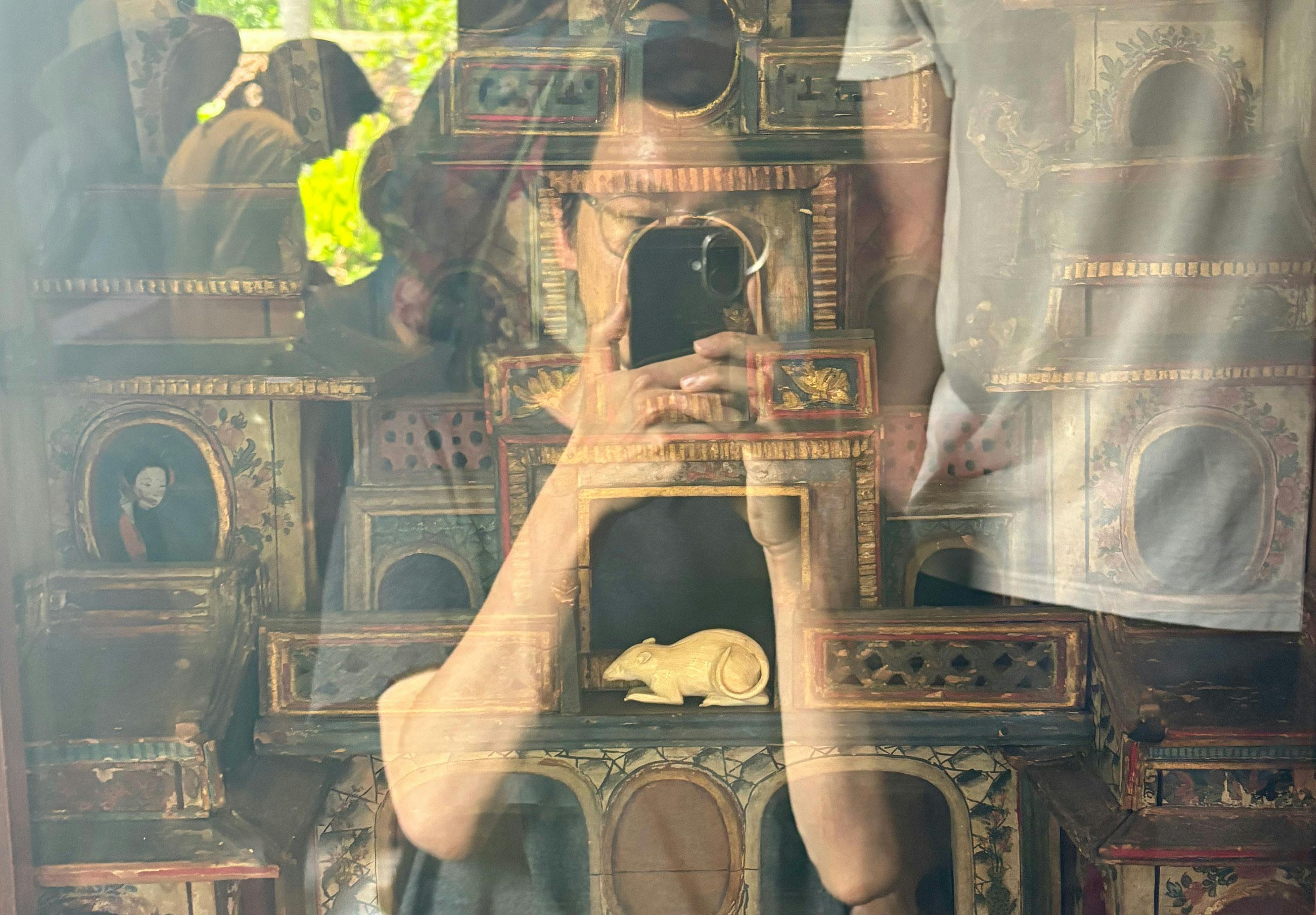 A person’s reflection holding a mobile appears on glass in front of a decorative box featuring a small statue of a white rat, painted portraits, and ornate details. Other reflections and greenery are visible in the background.