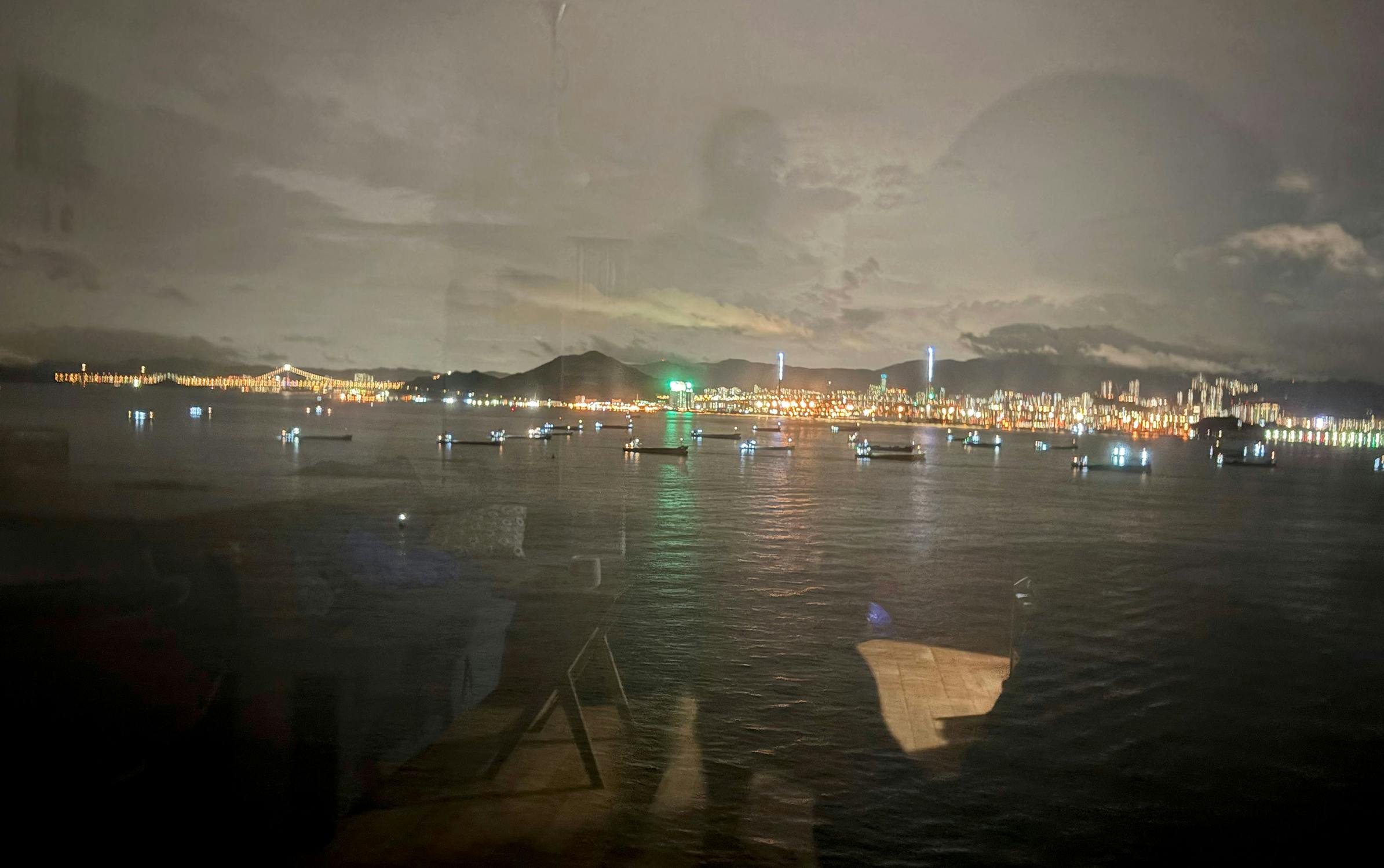 A night-time harbour scene with boats scattered on calm water, city lights glowing along the distant shoreline. The sky is cloudy, and faint reflections of indoor lights and silhouettes are visible on the window through which the photo was taken.