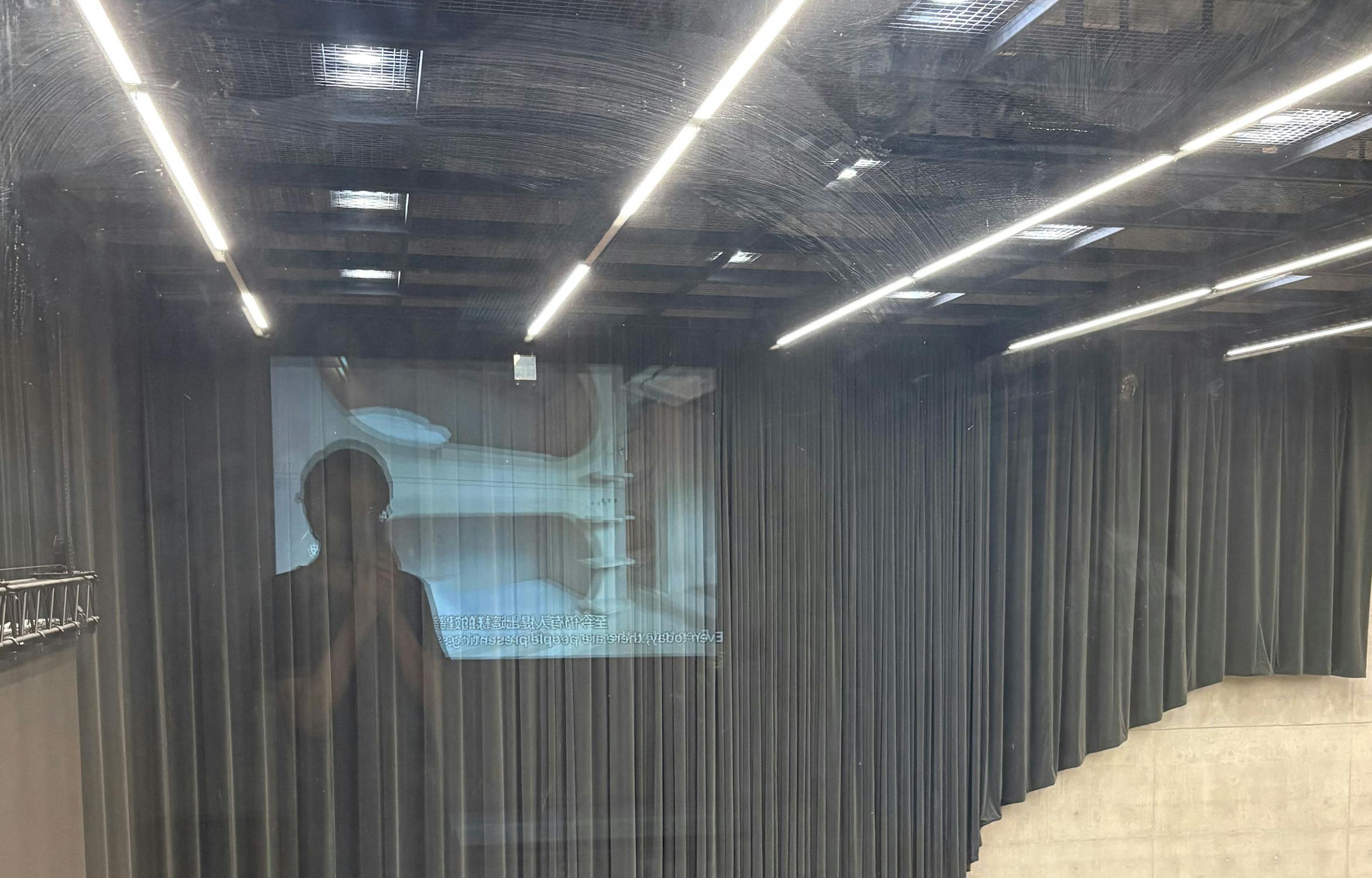Photo taken through a glass pane, reflecting ceiling lights and a person holding a mobile. Behind the glass, a dark curtain surrounds a projected image on the wall showing a double toilet and sink. The scene has an industrial, minimalist feel.