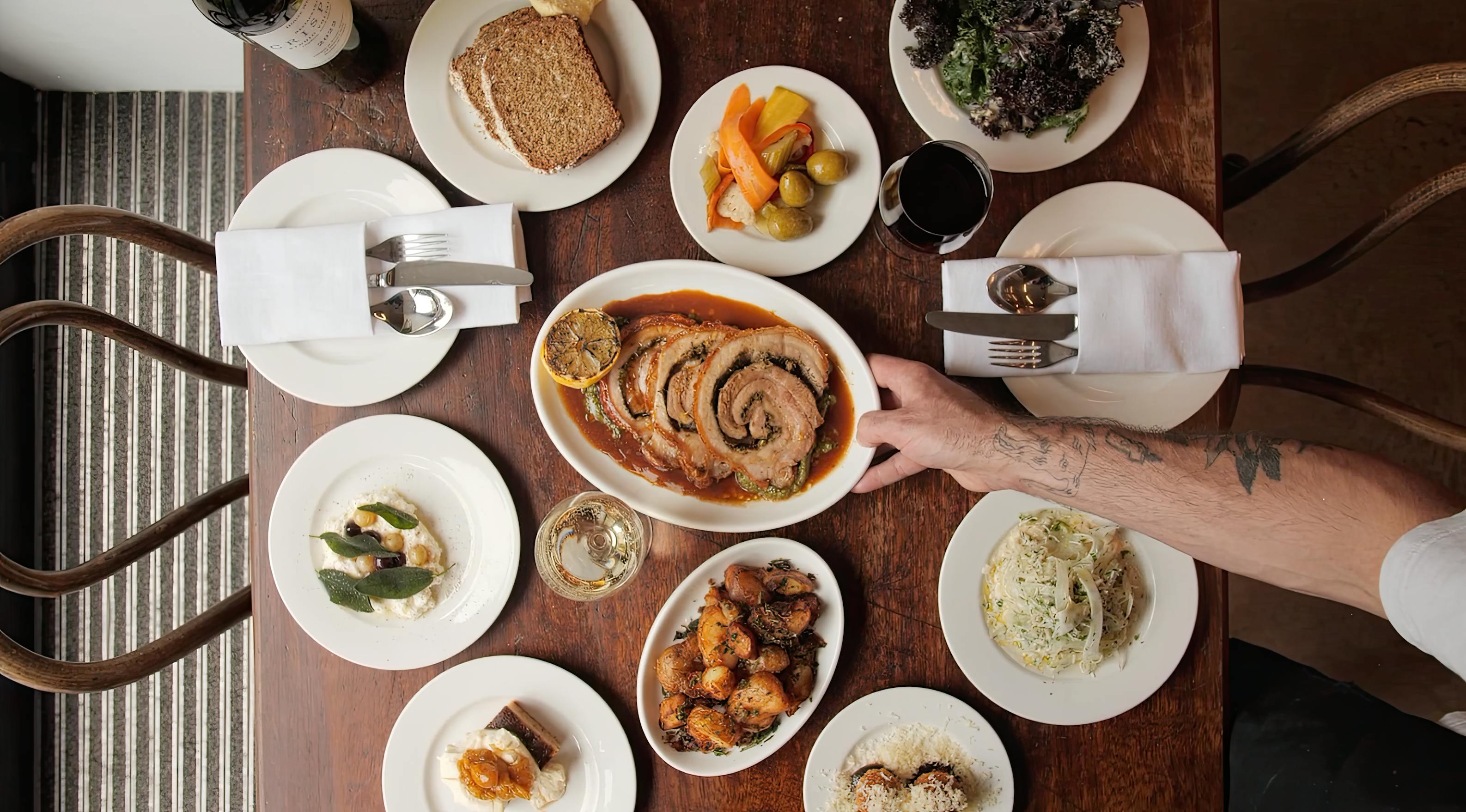 A wooden table laid with various dishes by Crispin at Studio Voltaire, including a central platter of rolled roasted meat with sauce, surrounded by plates of salad, vegetables, pasta, toast, roast potatoes, and puddings. A tattooed arm holds the meat platter; wine and water glasses are visible.