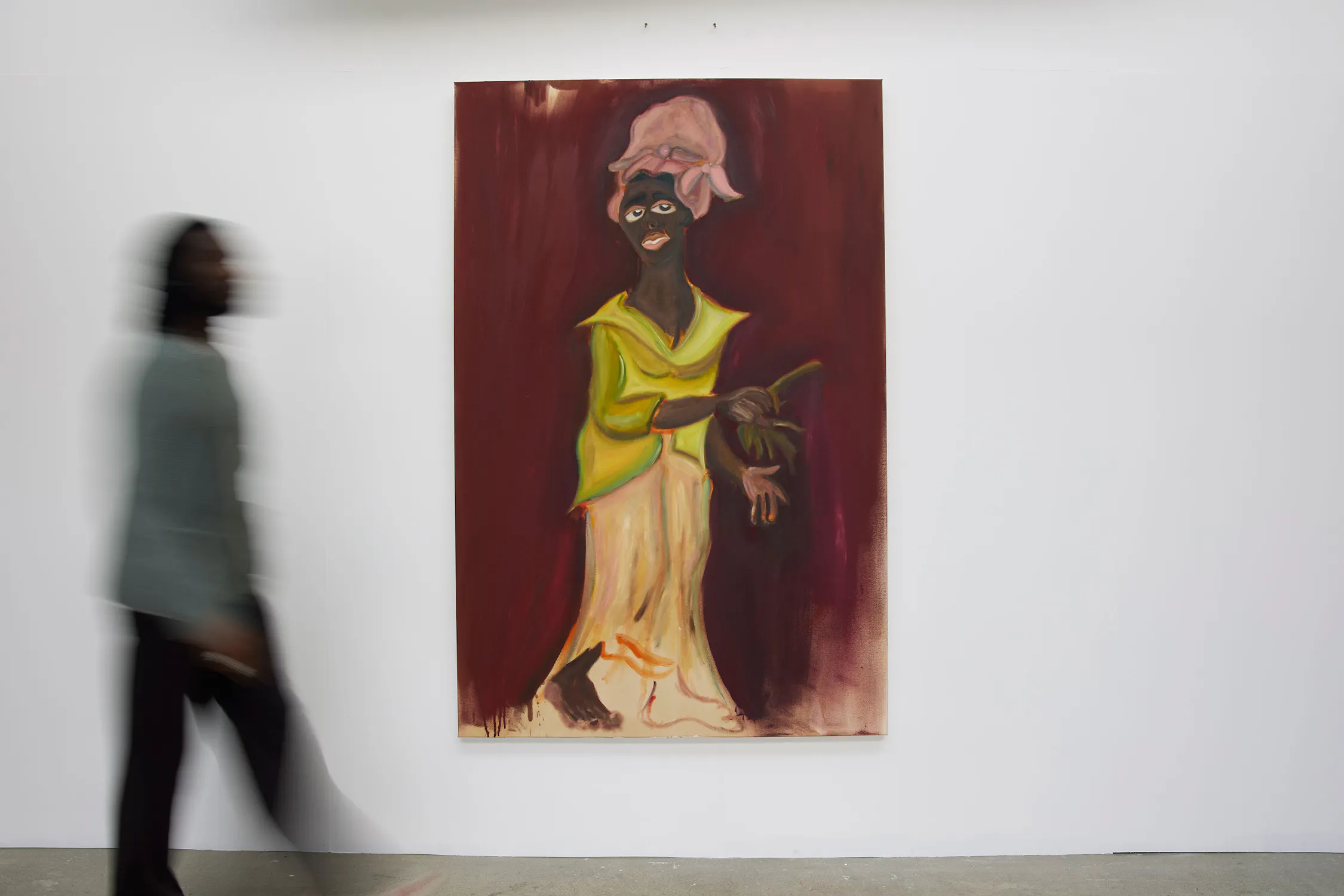 A person walks past a colourful painting on a white wall. The painting depicts a dark-skinned figure with exaggerated features, wearing a pink headwrap and yellow-green clothing, holding an object against a maroon background. The walker is blurred in motion.