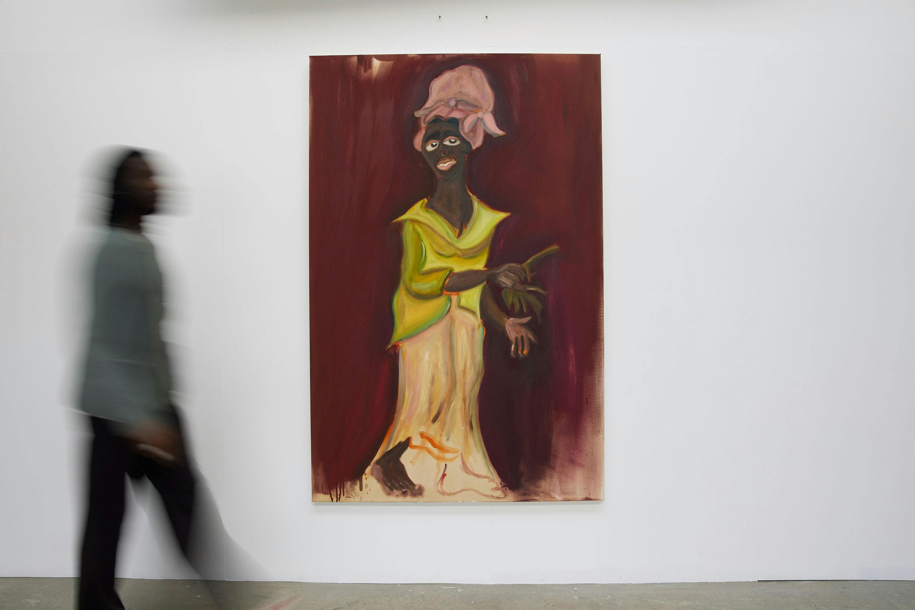 A person walks past a colourful painting on a white wall. The painting depicts a dark-skinned figure with exaggerated features, wearing a pink headwrap and yellow-green clothing, holding an object against a maroon background. The walker is blurred in motion.