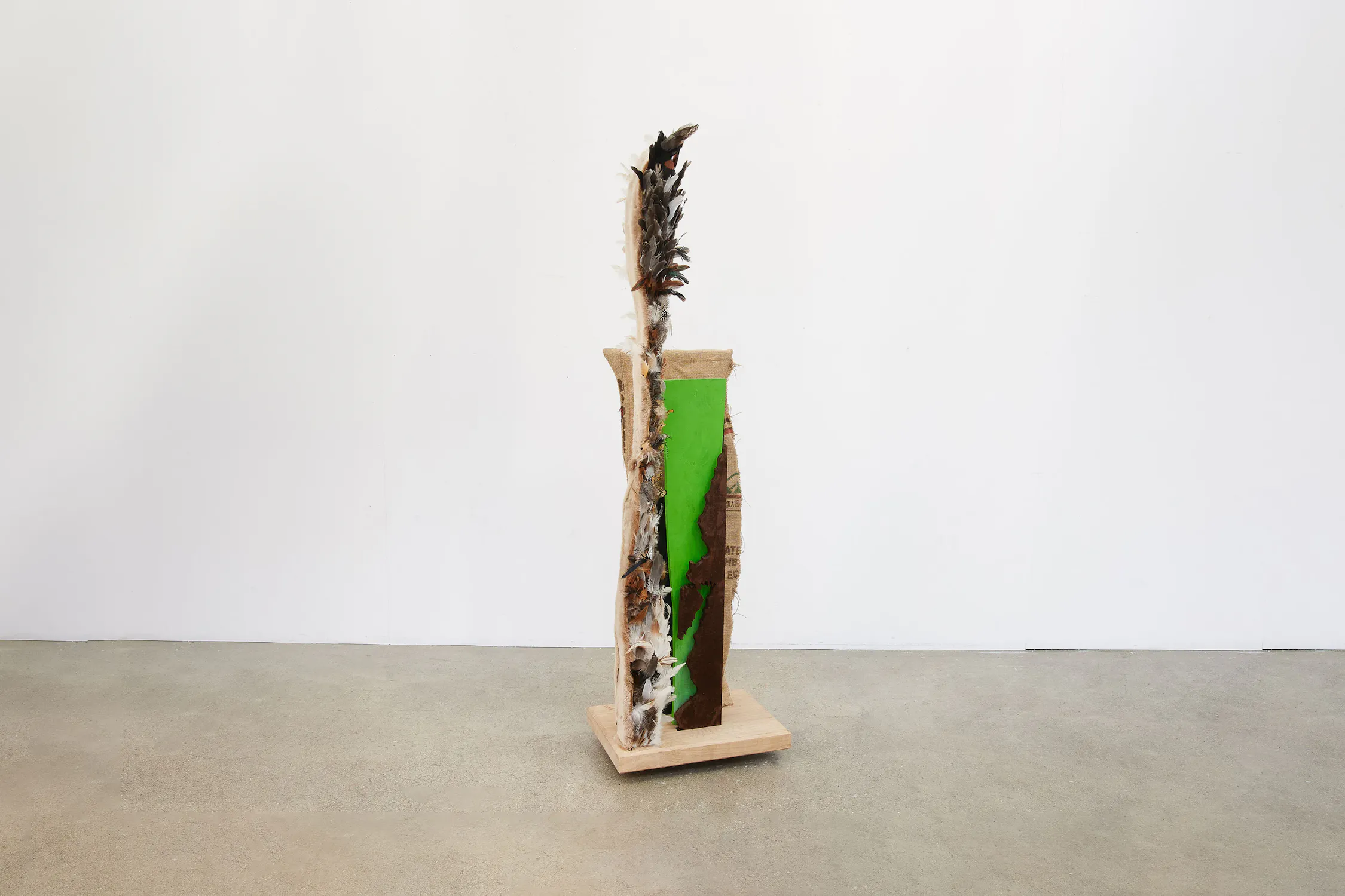 A vertical mixed-media sculpture stands on a wooden base, featuring layered textures of fabric, cardboard, and paint. The front shows a bright green panel with a brown shape, whilst rough, organic materials and a feathered element extend from the top.