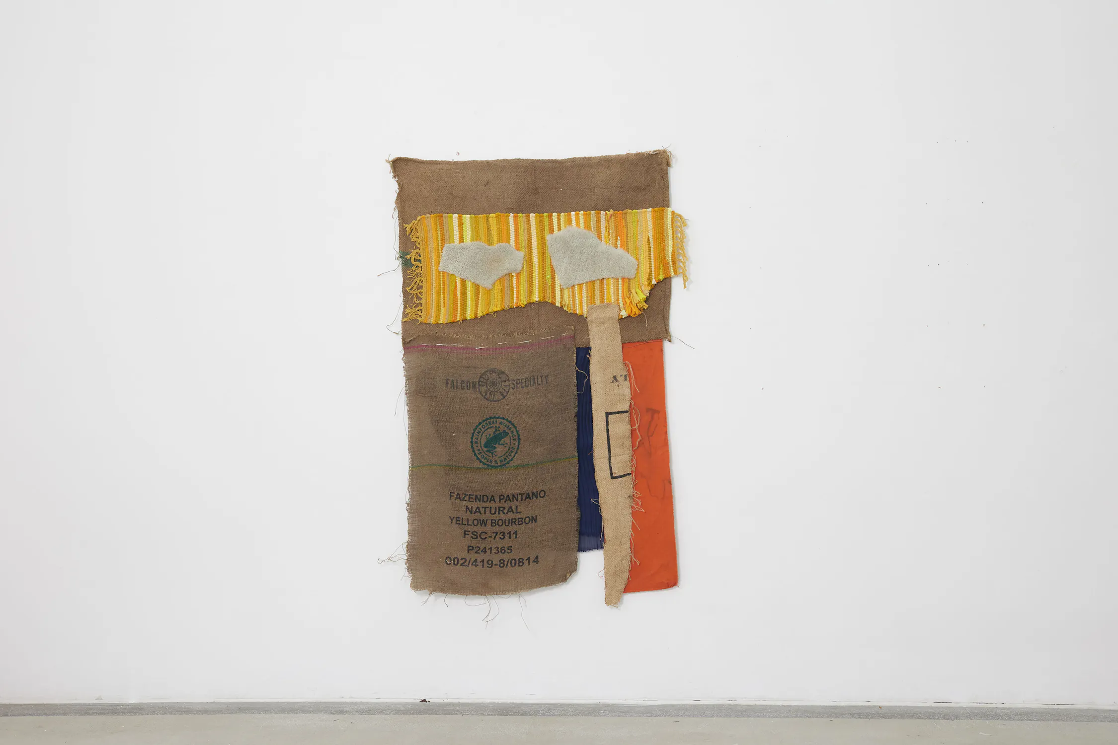 A hanging hessian sack with rough edges is mounted on a white wall. The sack has a printed green label at the top and a rectangular patch of bark in the centre. A brown bandana-like cloth peeks out from the bottom. The floor is grey concrete.