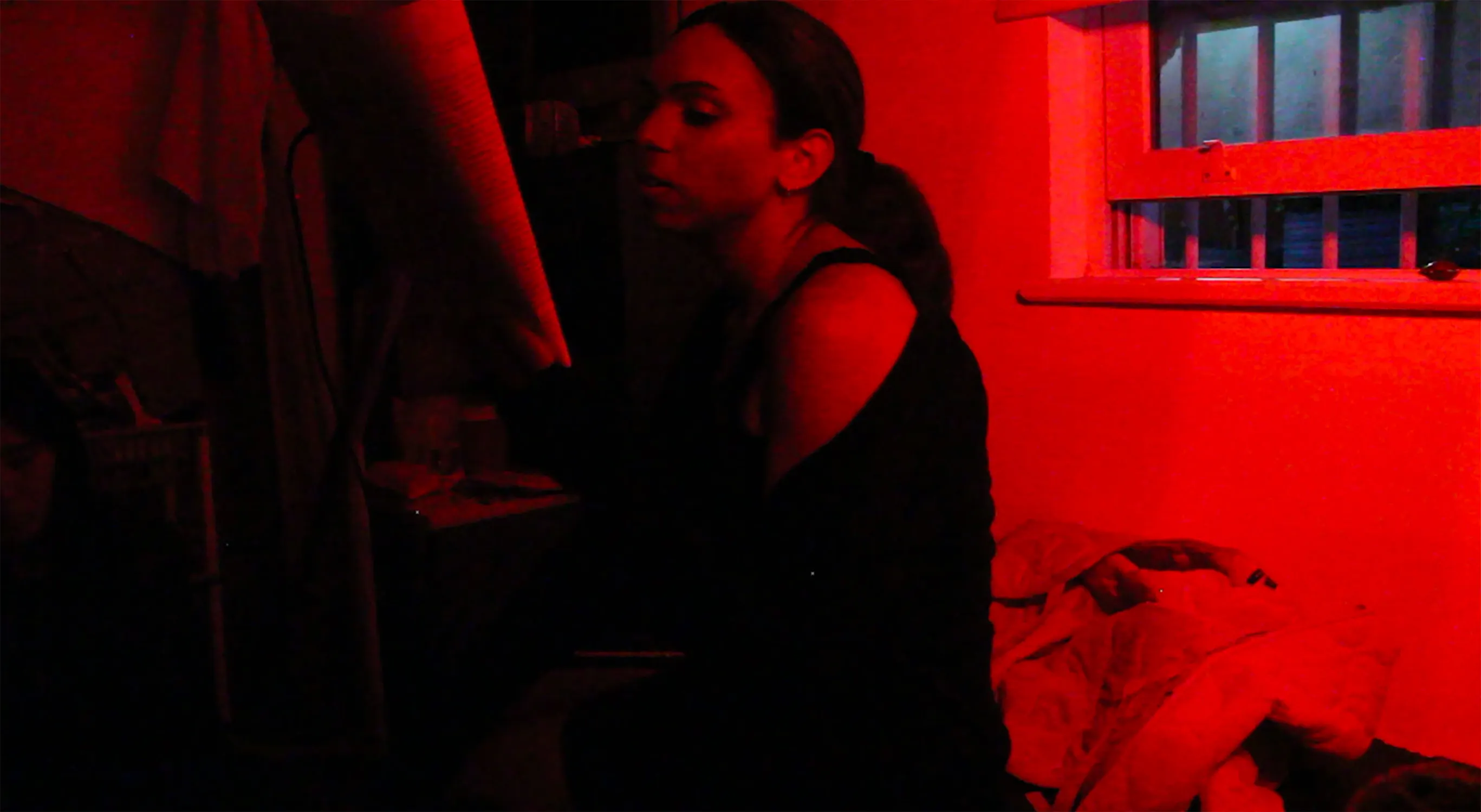 A person with long dark hair, wearing a black off-the-shoulder top, sits on an untidy bed in a dimly lit room bathed in red light. A window with white blinds and various objects are visible in the background. The atmosphere appears tense or contemplative.