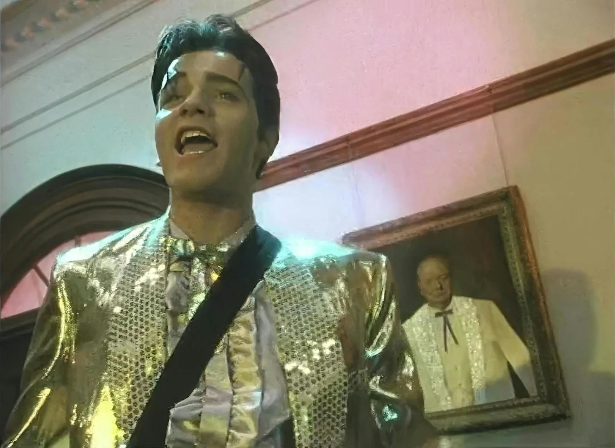 A man in a shiny gold and silver suit sings passionately indoors, wearing an eye patch and shoulder sash. Behind him, a framed portrait of an older man in formal attire hangs on a wall lit with pink and green light.