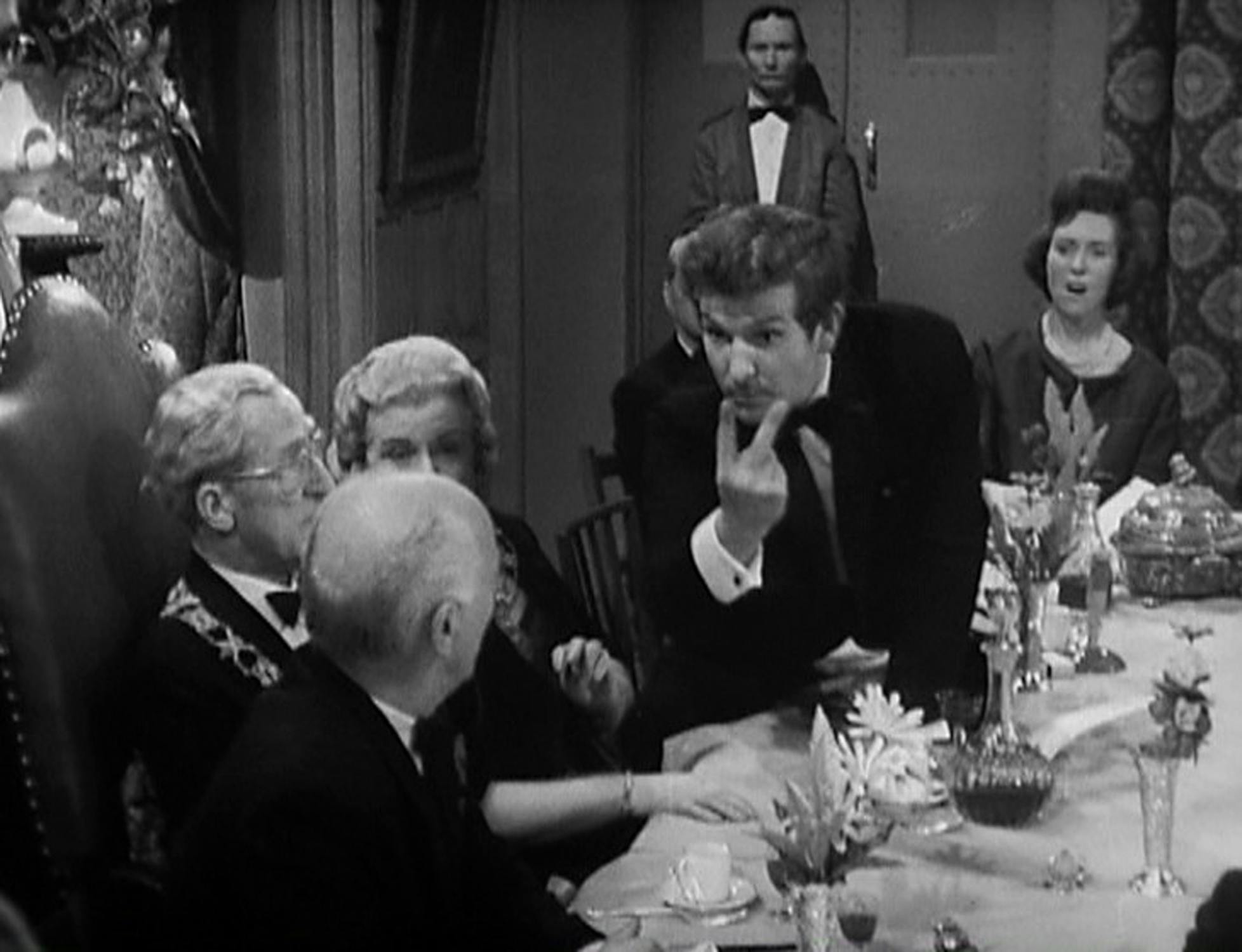 A black-and-white image shows a lively dinner scene. A man in a suit gestures with two fingers at a formal table where several adults are seated, watching him. The table is laid with glasses, flowers, and a decanter. A man stands in the background.