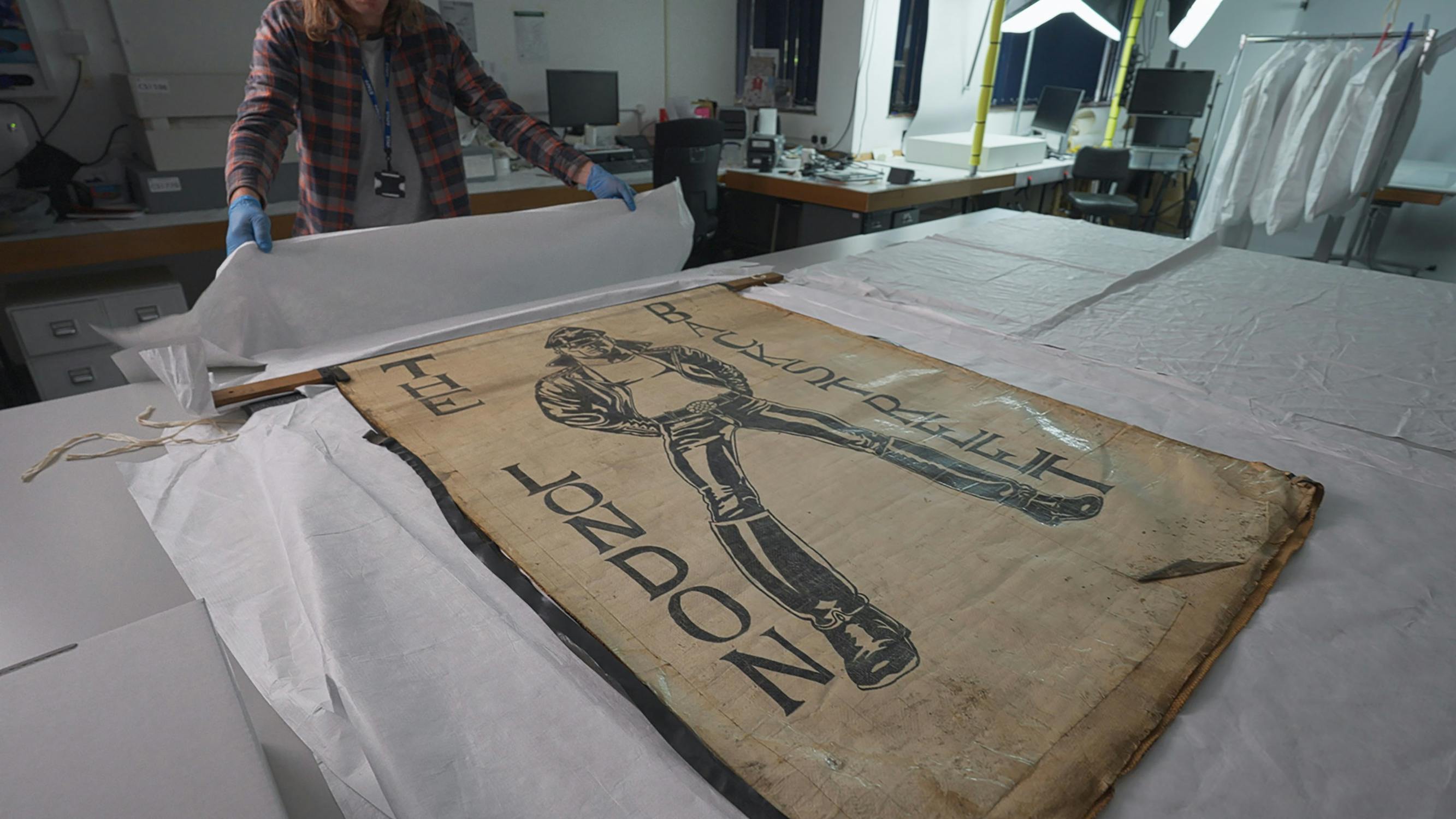 A person wearing blue gloves and a checked shirt carefully wraps white tissue paper around an old, illustrated fabric banner labelled “THE LONDON BACKSTREET.” The banner shows a person in tall boots. The scene is in a conservation or archival workspace.
