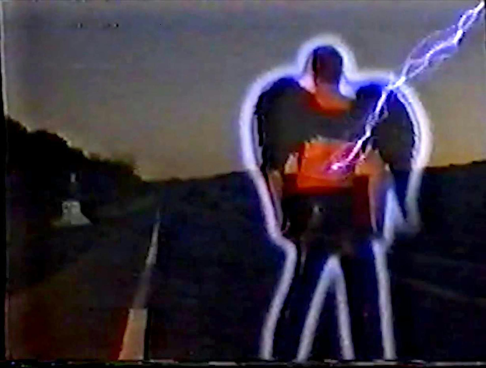 A person outlined in glowing white light stands on a dark, empty road at dusk. A jagged bolt of blue lightning strikes their chest, creating a vivid electric effect. Trees and a distant horizon are visible against the dim sky. The image has a grainy, retro look.