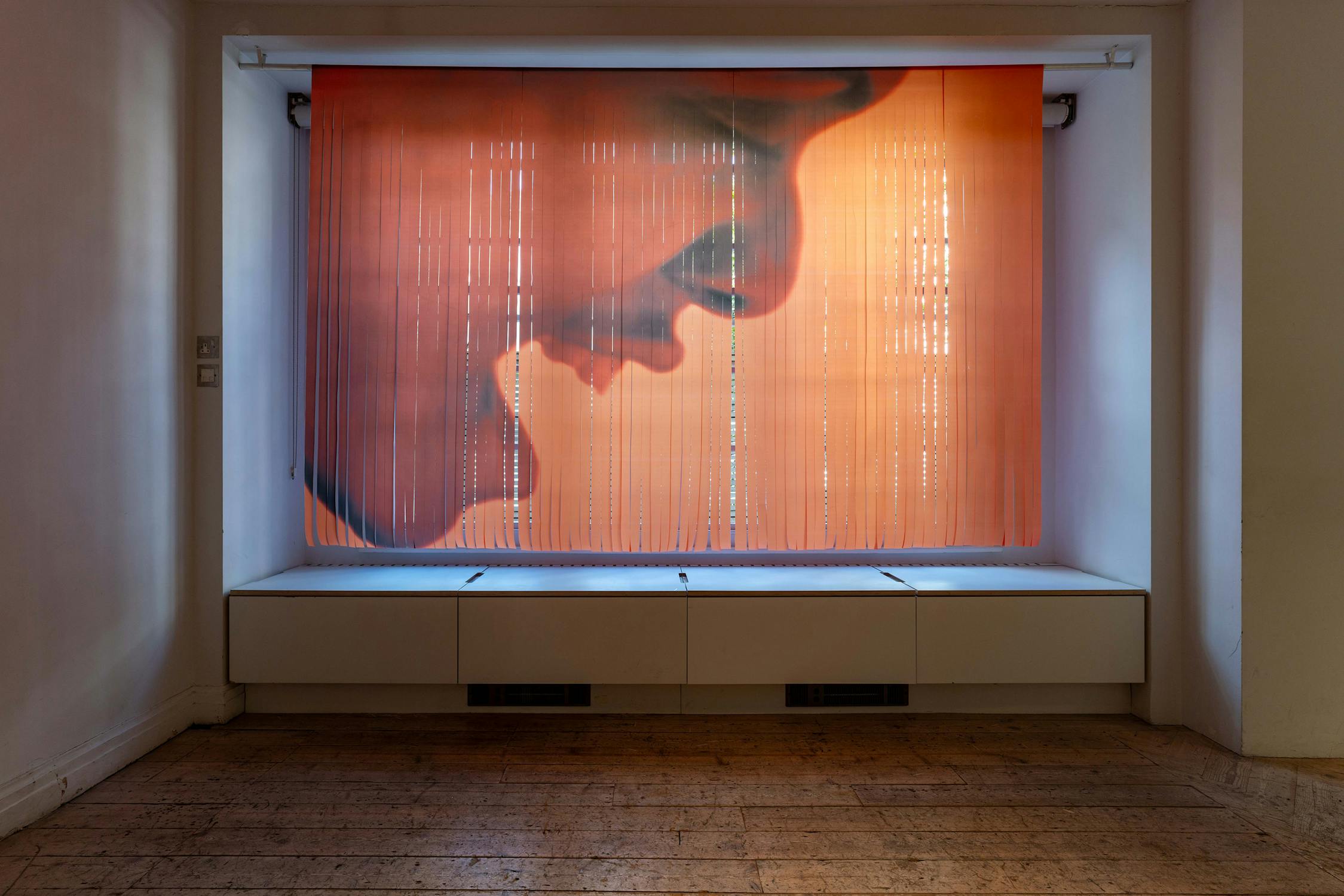 A translucent curtain with a large, side-profile image of a person's face in warm orange tones hangs in a recessed window alcove with white walls and built-in storage drawers below, on a wooden floor in an otherwise empty room.