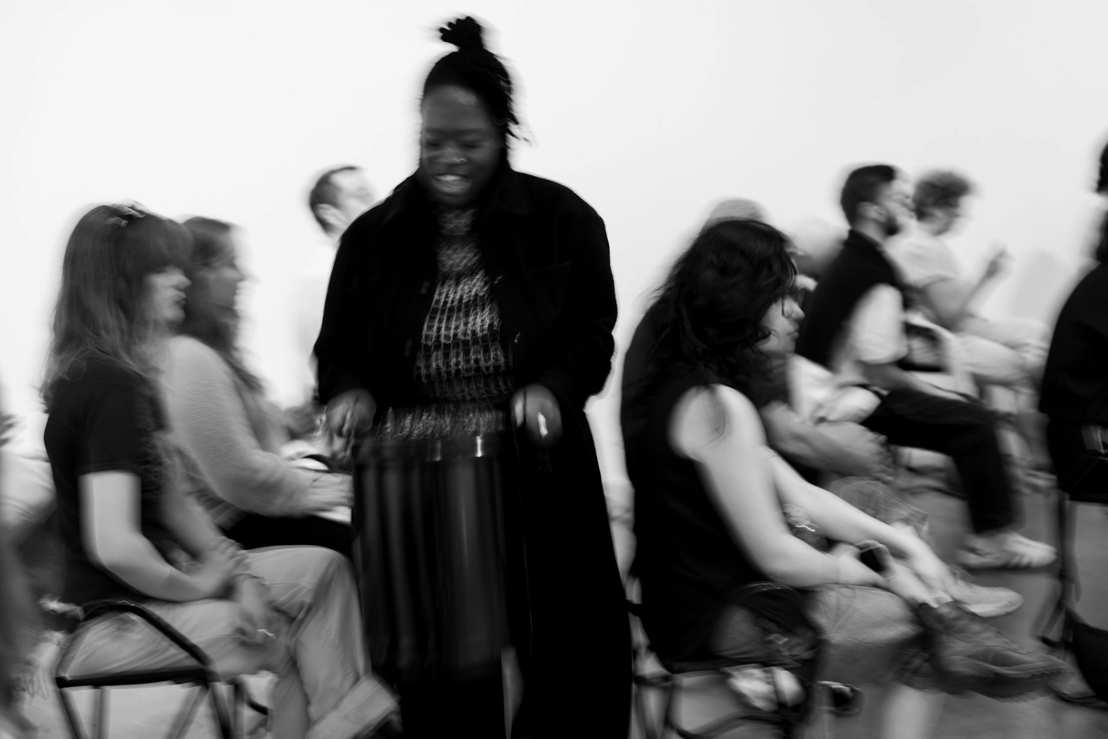 Black and white photo of people seated in rows, some blurred in motion. A person stands in the centre with hands on a drum, appearing to play it. The background is minimal, with other seated people facing sideways and wearing casual clothes. The mood is dynamic and lively.