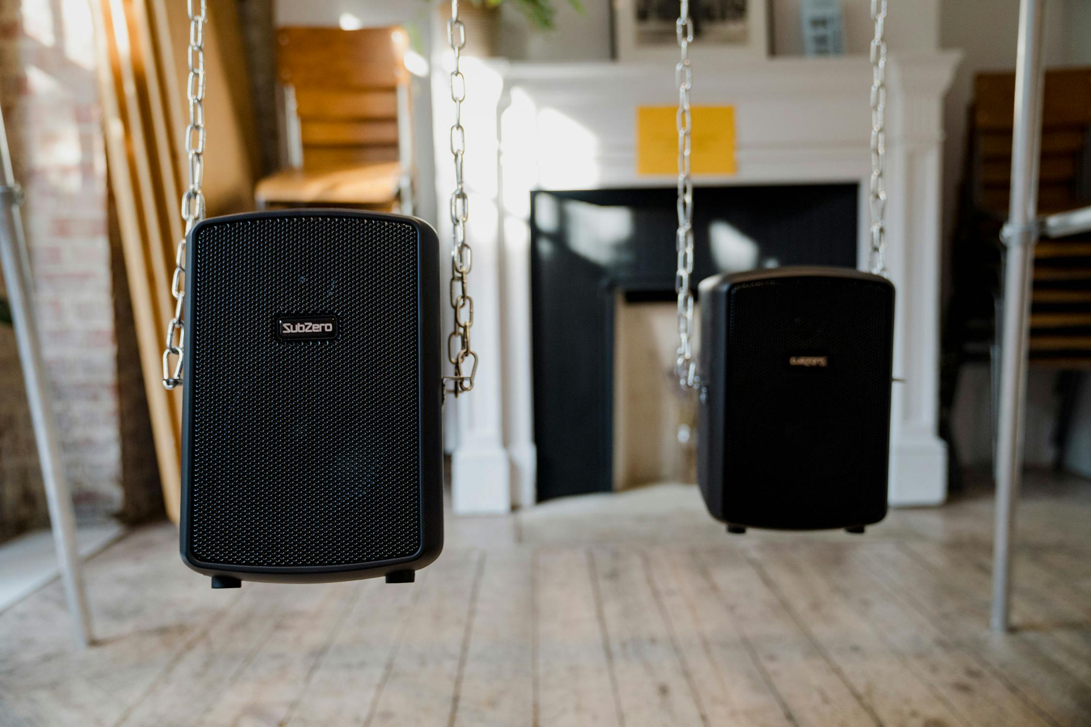 Two black SubZero speakers hang from metal chains in a bright room with wooden floors, white panelled walls, and a fireplace in the background. Sunlight streams in from the left, highlighting the left speaker and part of the floor.