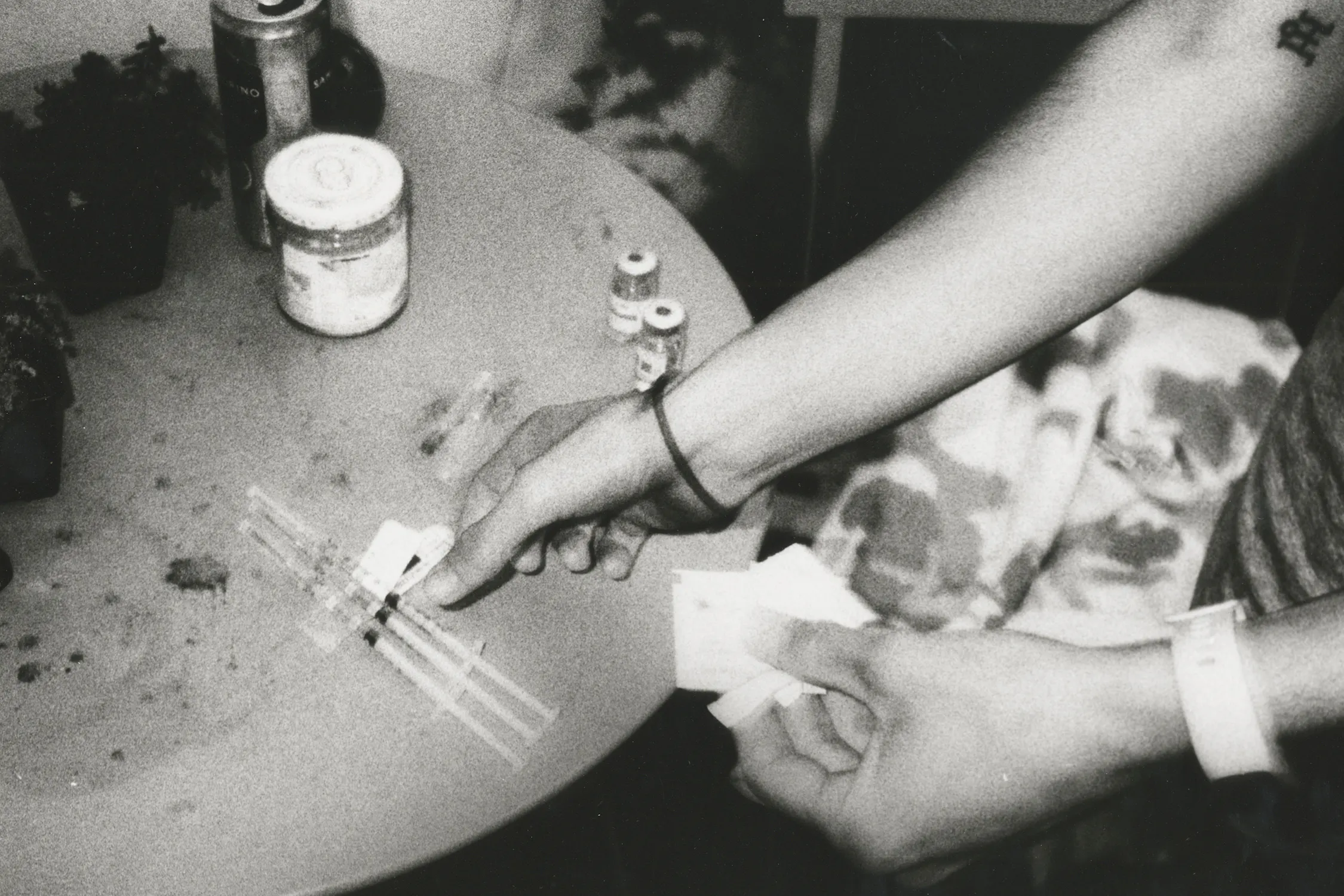 A black and white photo of a person's hands preparing testosterone shots on a cluttered round table. One hand holds paper packets, the other handles syringes and small vials. Scattered items and bottles are visible, suggesting a cluttered, tense environment.