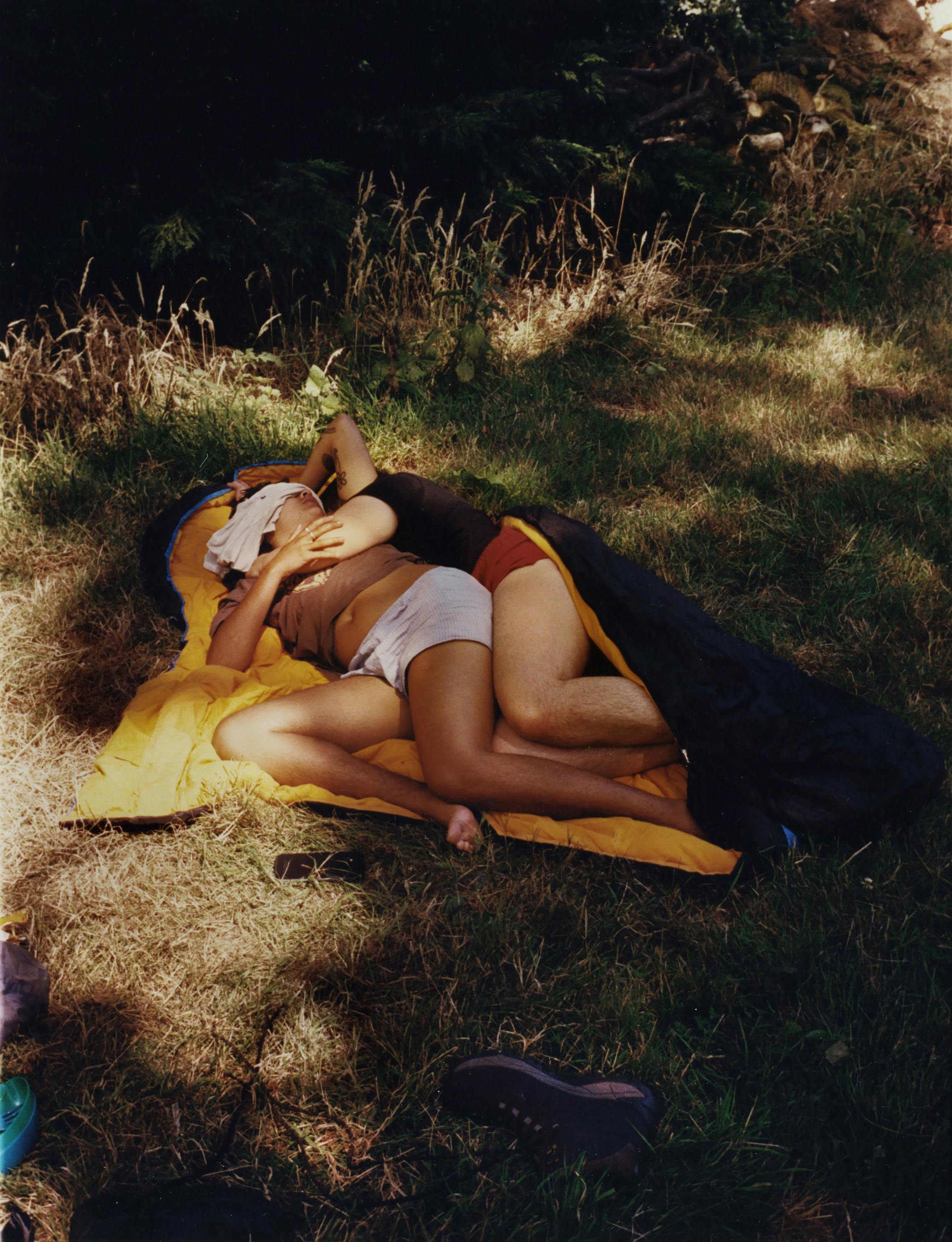 Two people lie closely together on a yellow sleeping mat outdoors, partly covered by a dark blanket. They are embracing each other in a grassy, sun-dappled area, surrounded by trees and shadows, creating an intimate and relaxed atmosphere.